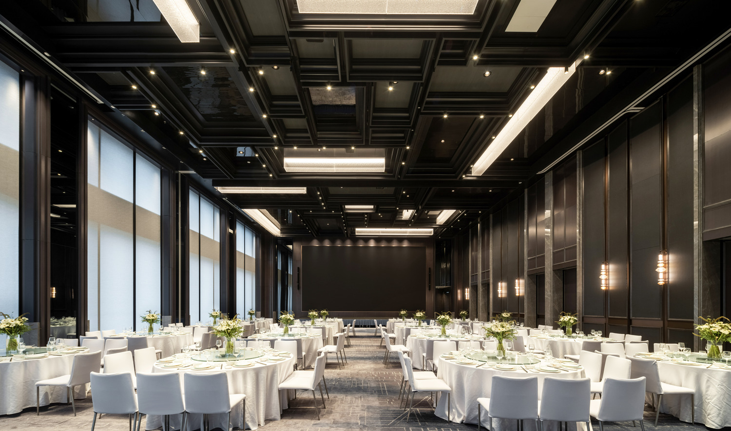 Spacious hotel ballroom hall with round tables set for an event. Each table has white tablecloths, green floral centerpieces, and elegant place settings.