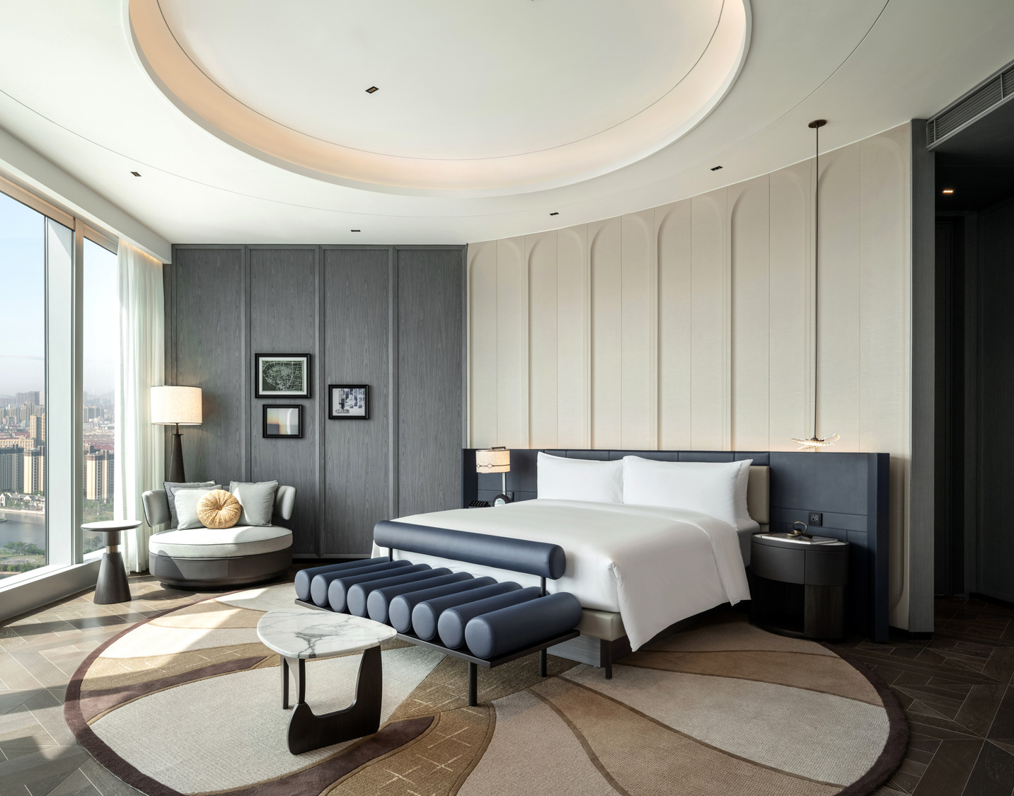 Elegant bedroom with neutral tones. Features a large bed with black pillows, flanked by bedside lamps. A plush bench and round table add a cozy touch.