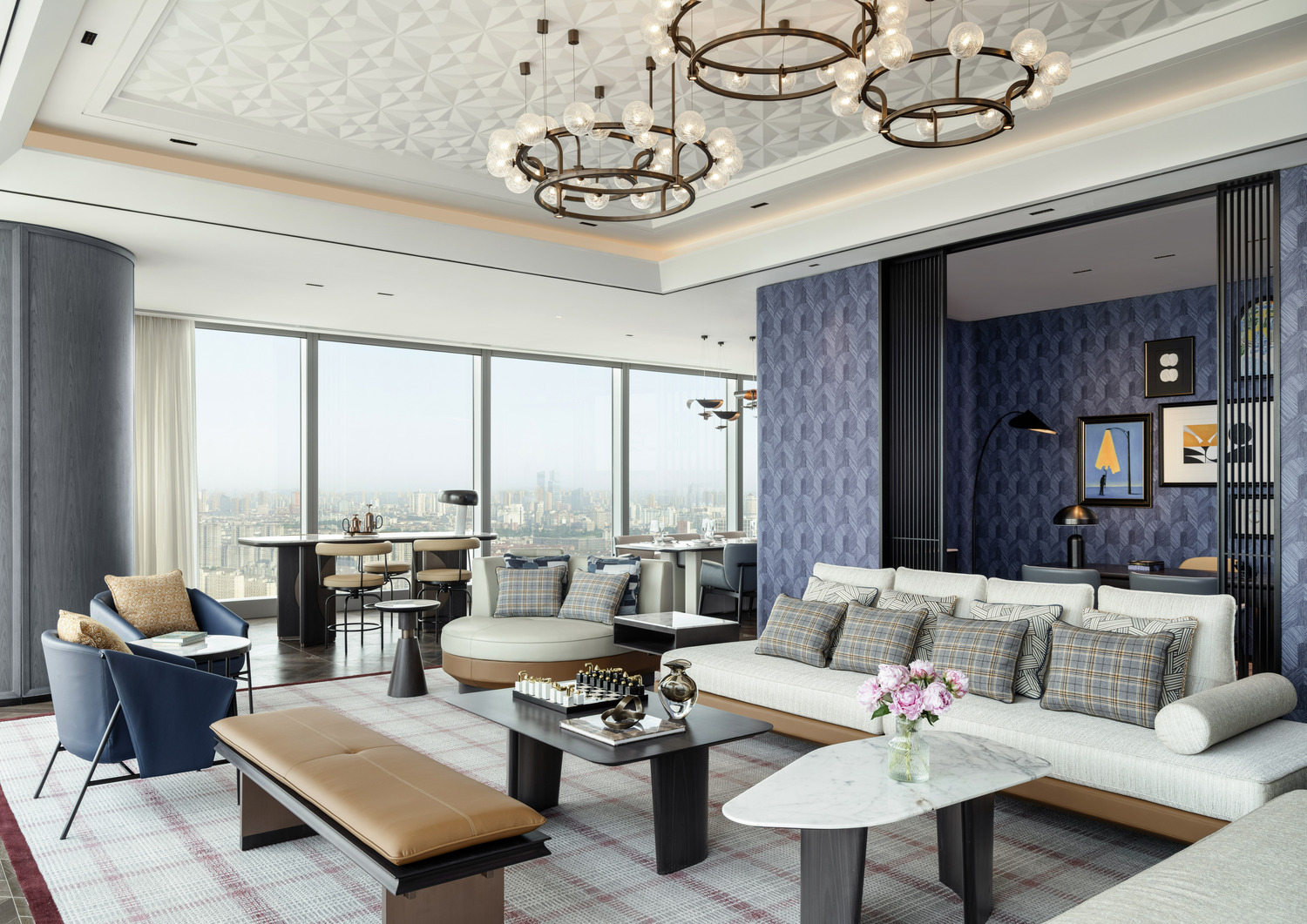Elegant bedroom with neutral tones. Features a large bed with black pillows, flanked by bedside lamps. A plush bench and round table add a cozy touch.