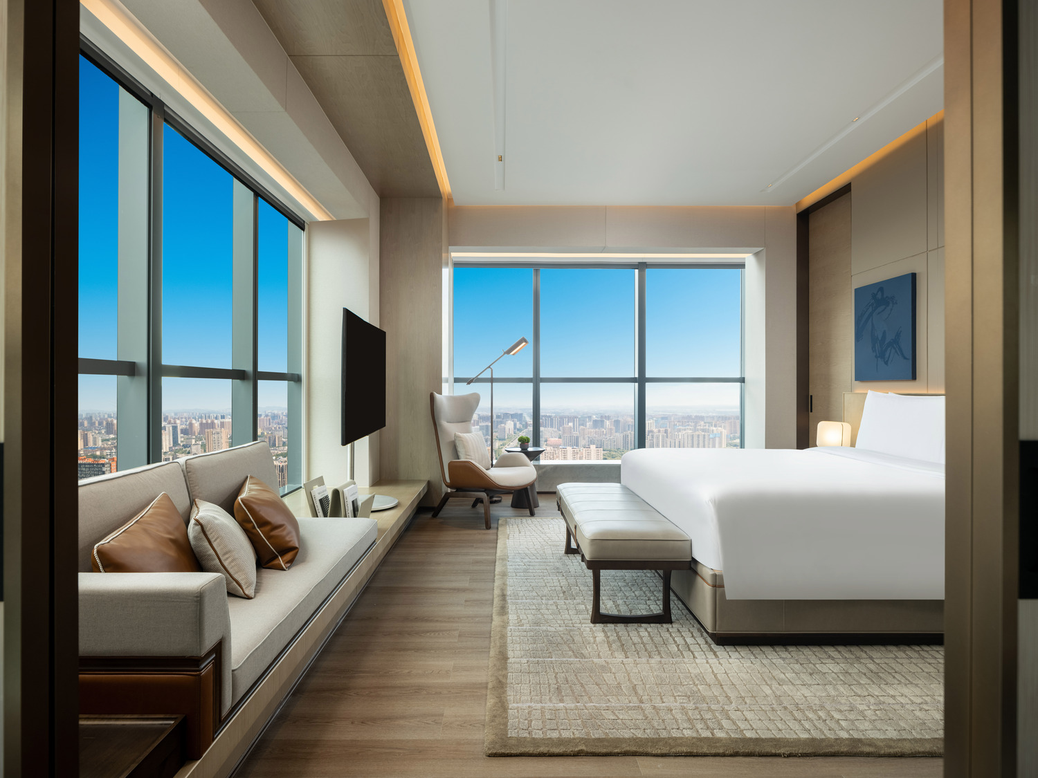 Elegant bedroom with neutral tones. Features a large bed with black pillows, flanked by bedside lamps. A plush bench and round table add a cozy touch.