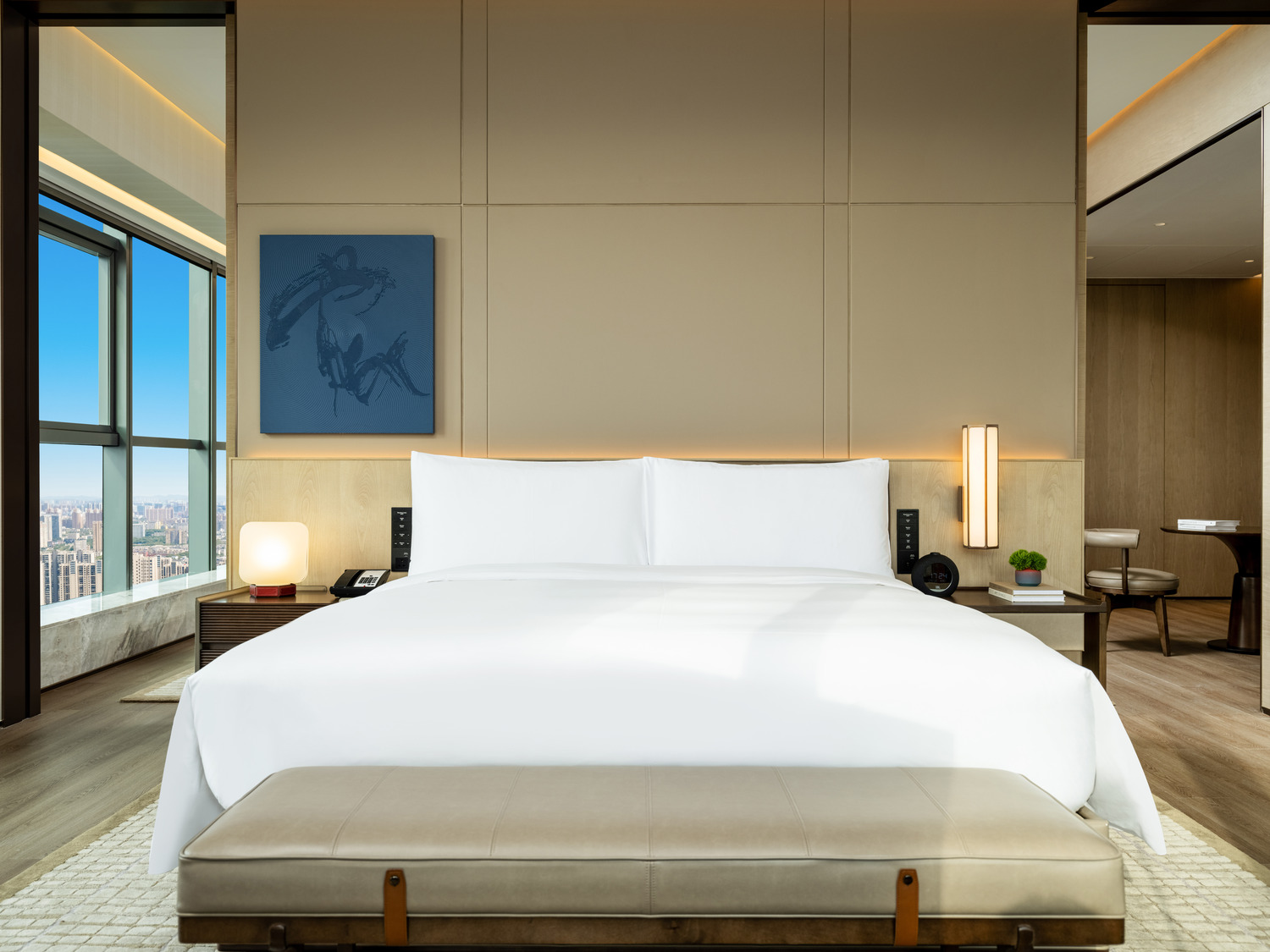 Elegant bedroom with neutral tones. Features a large bed with black pillows, flanked by bedside lamps. A plush bench and round table add a cozy touch.