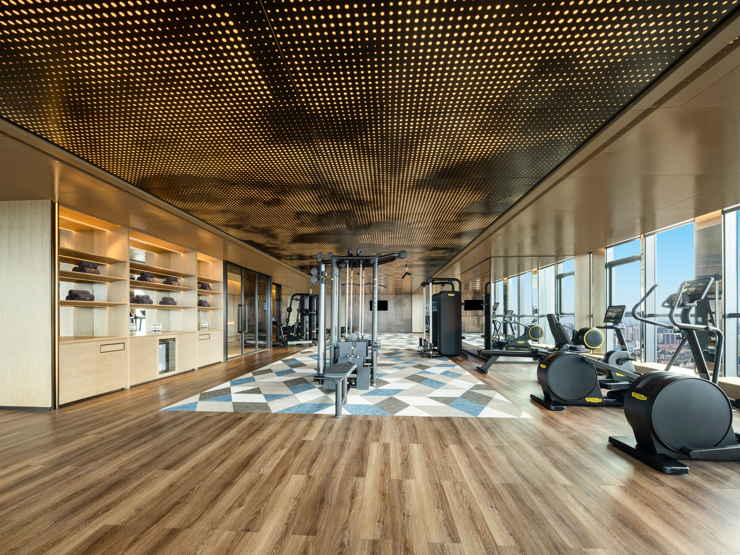 Spacious, modern gym with a wooden ceiling and large windows overlooking a cityscape. Equipped with treadmills, exercise bikes, and benches. Luxurious ambiance.
