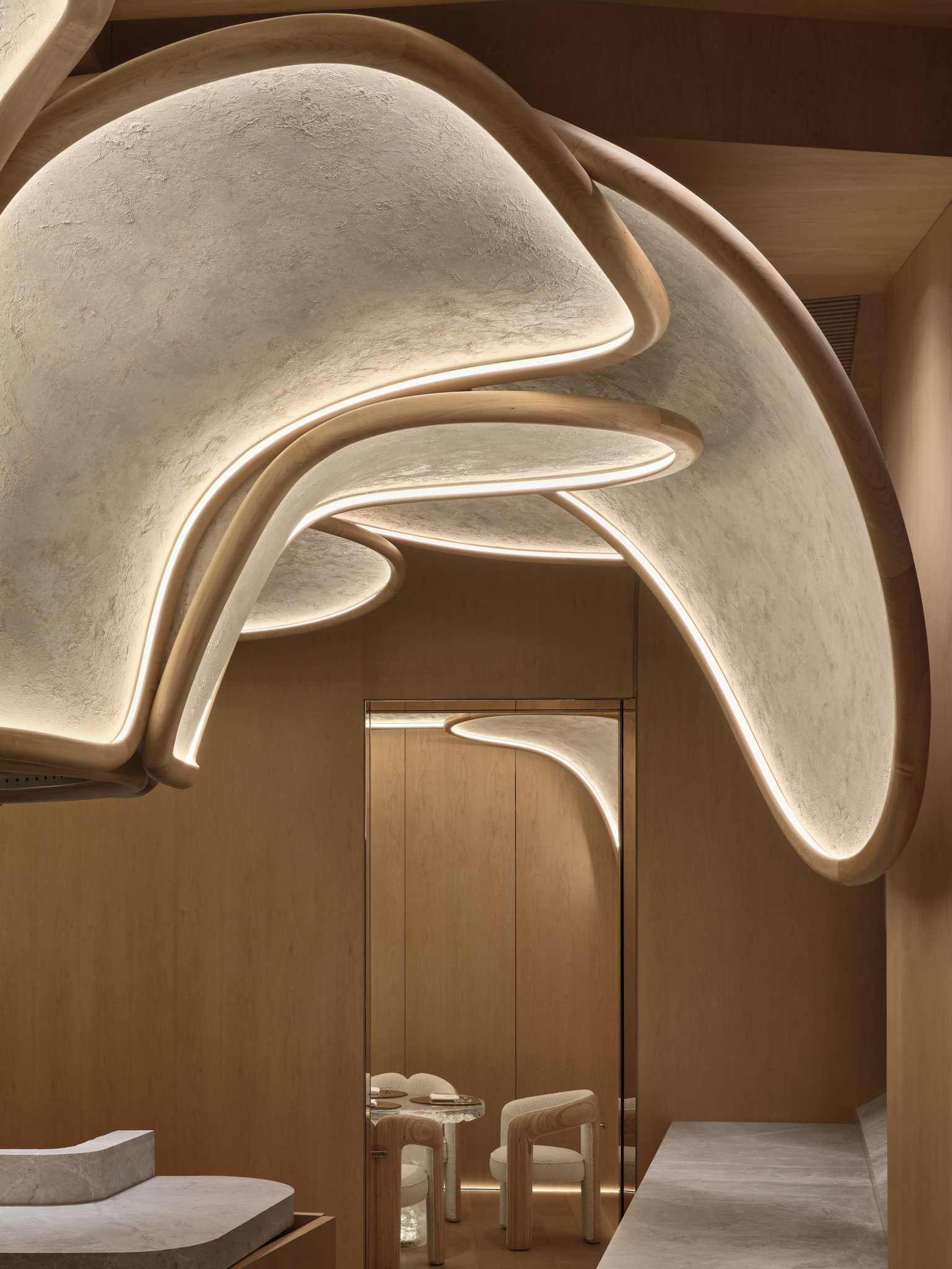 Delicate curved, petal-like ceiling design with soft lights and a sleek mirror wall, creating an elegant, serene ambiance. Light wood tones dominate.