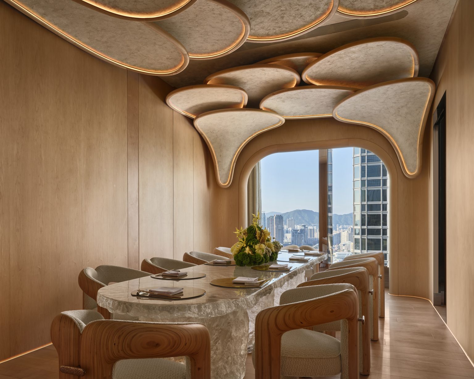 Elegant dining room with wavy ceiling panels, a long marble table, modern wooden chairs, and a large window showcasing a city skyline view.