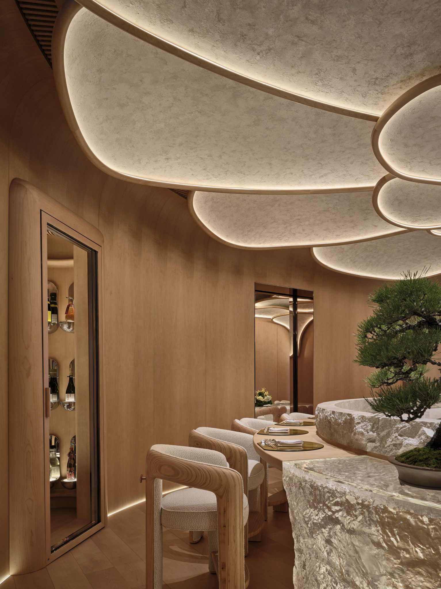 4. Elegant dining room with soft wooden tones. A glass cabinet displays various bottles. Curved chairs and table evoke a serene, modern ambiance.
