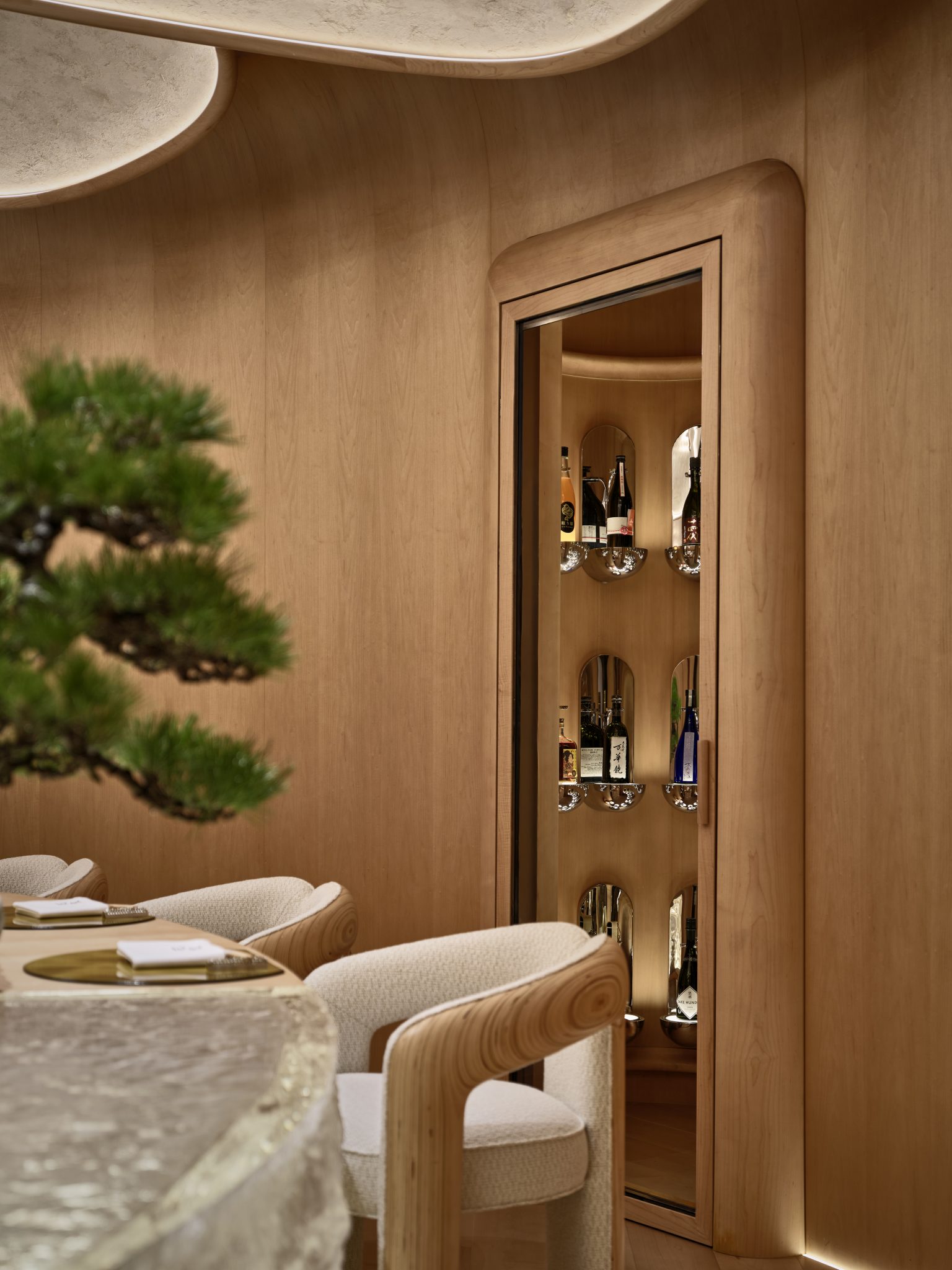 4. Elegant dining room with soft wooden tones. A glass cabinet displays various bottles. Curved chairs and table evoke a serene, modern ambiance. a small green bonsai peeked through.