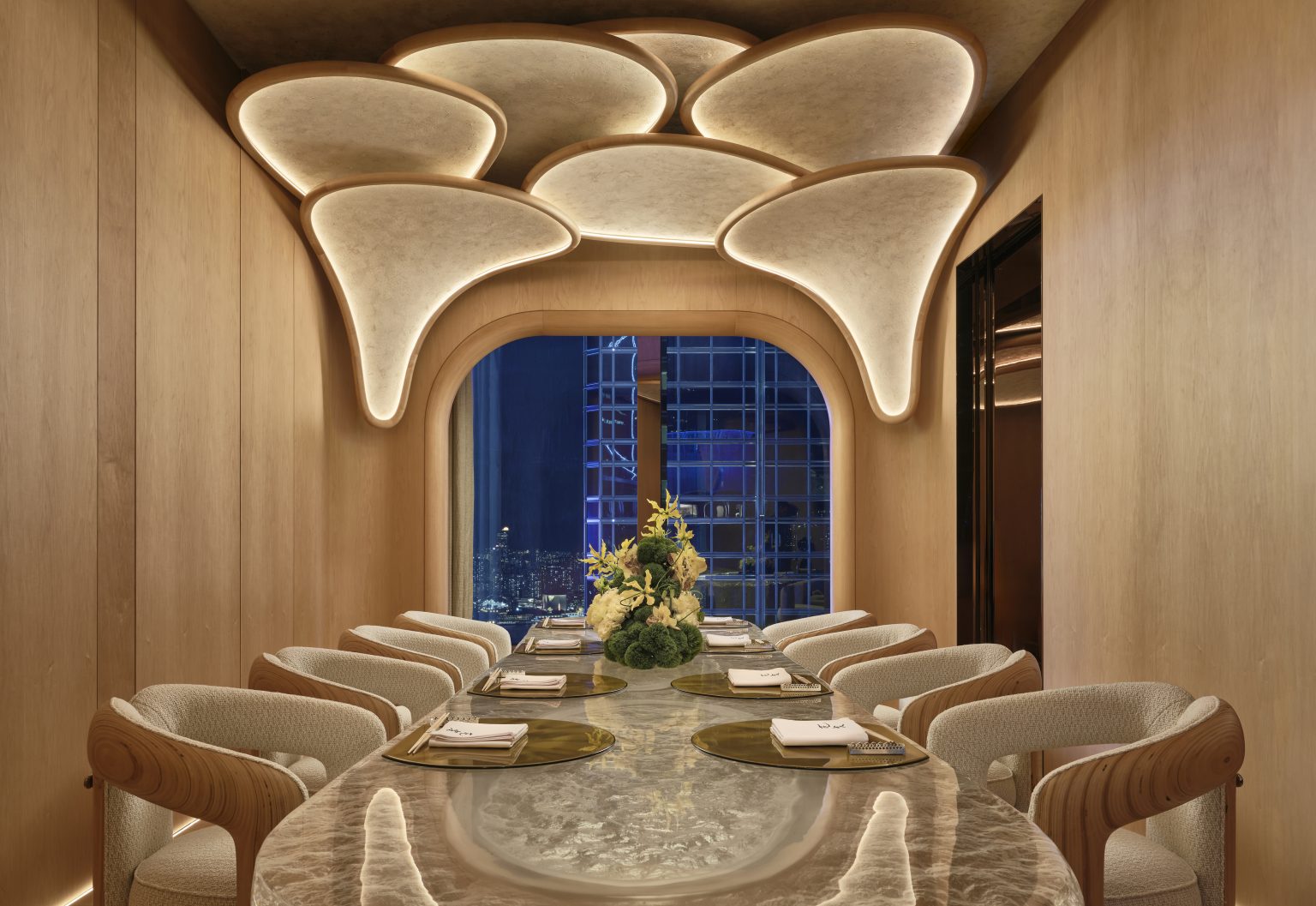 Luxurious private dining room with a polished marble table, elegant beige chairs, and soft curved ceiling lights. A cityscape view is visible through a large window.