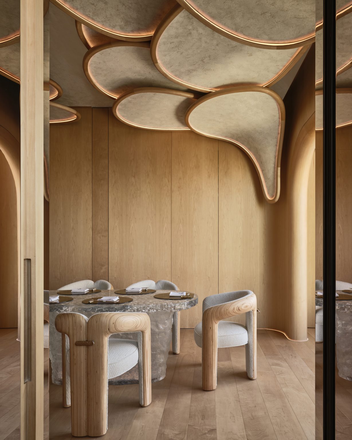 Warm, modern dining room with a natural wooden theme. Features sculptural ceiling designs, curved wooden walls, and sleek chairs around a polished table.