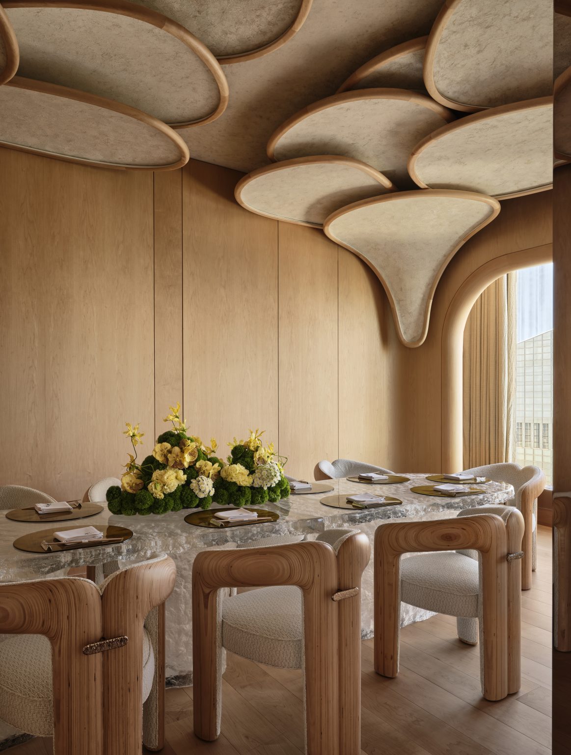 Wood-paneled dining room with sculptural beige ceiling. Curved wooden chairs surround a marble table with a lush yellow and white flower centerpiece.