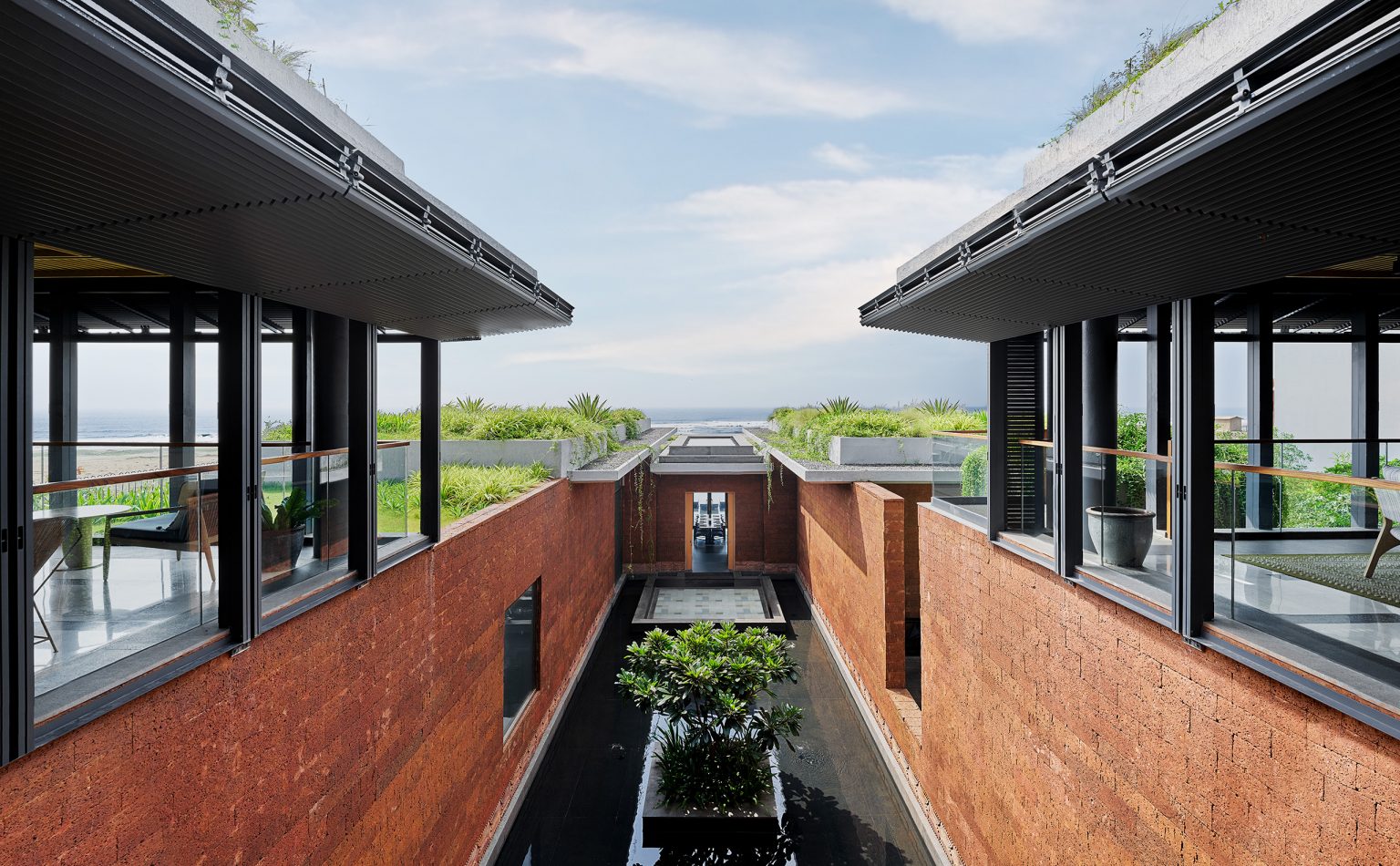 Modern, open-air building with red brick walls and water feature. Green plants line the walkway. Sky and ocean visible in the background, conveying tranquility.