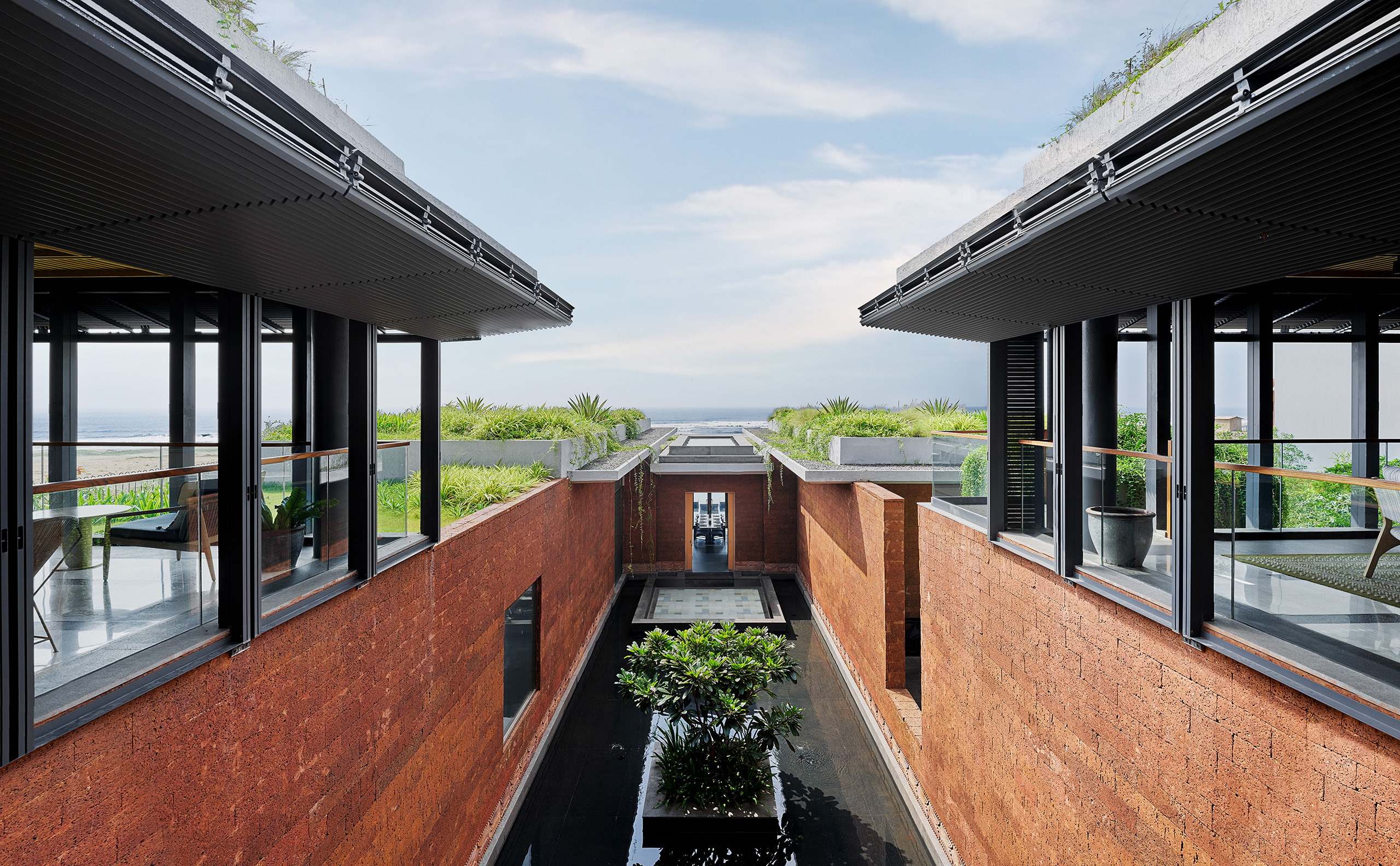Modern, open-air building with red brick walls and water feature. Green plants line the walkway. Sky and ocean visible in the background, conveying tranquility.