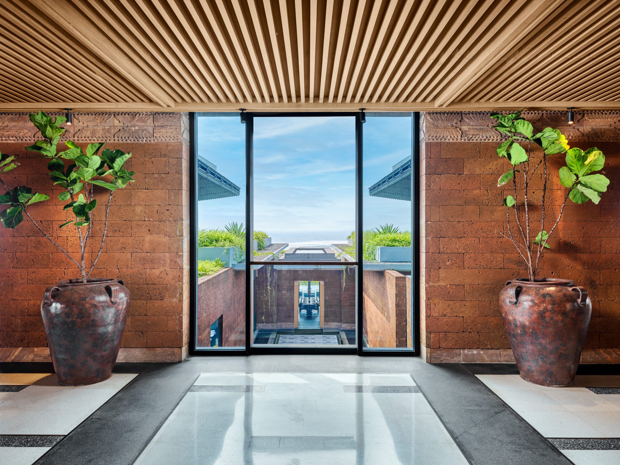 Spacious modern hallway with large potted plants on each side, featuring a floor-to-ceiling glass window overlooking a scenic garden and ocean view.