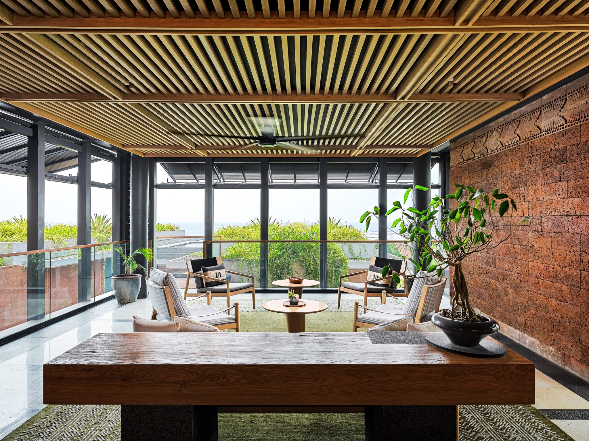 A serene lounge with modern decor features wood-paneled ceilings, a potted plant, and seating around a round table, with large windows offering a lush view.