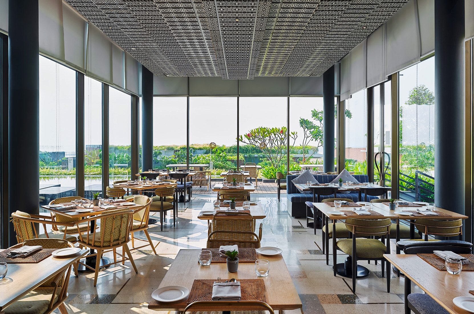 Spacious restaurant with wooden tables and wicker chairs, surrounded by large glass windows offering a view of lush greenery, creating a serene atmosphere.