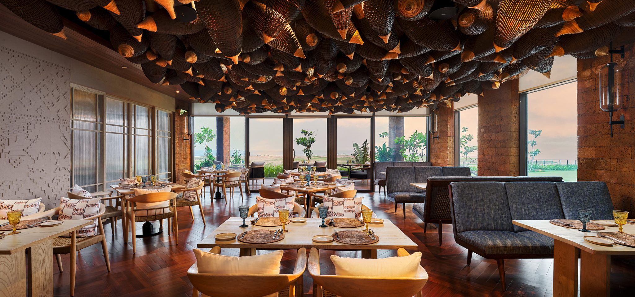 Warm, elegant restaurant interior with wooden tables, cozy chairs, and decorative ceiling lights. Large windows reveal a scenic outdoor view.