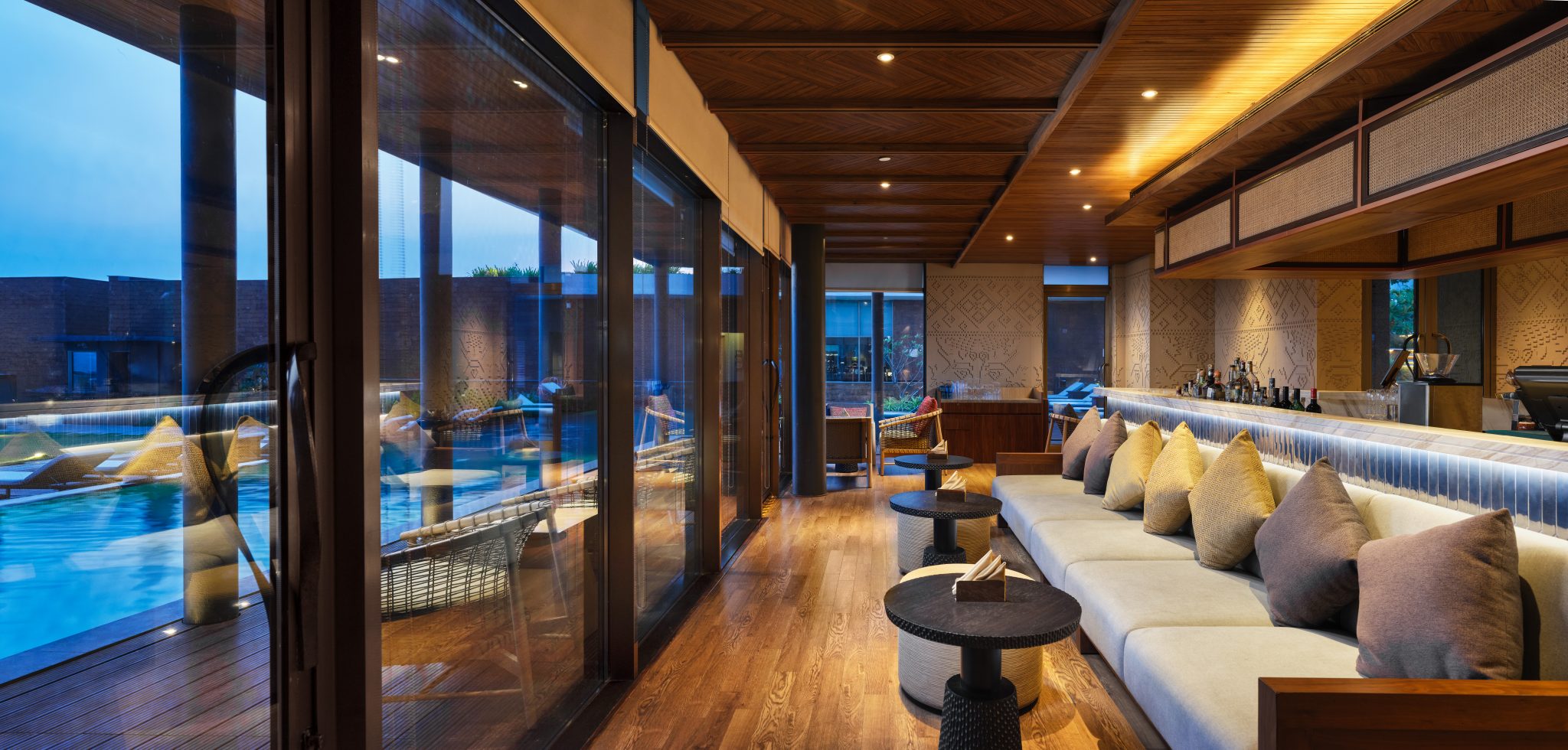 Modern lounge with wooden floors, plush beige sofas, and numerous pillows, beside a large window showing a pool. Warm lighting creates a cozy ambiance.