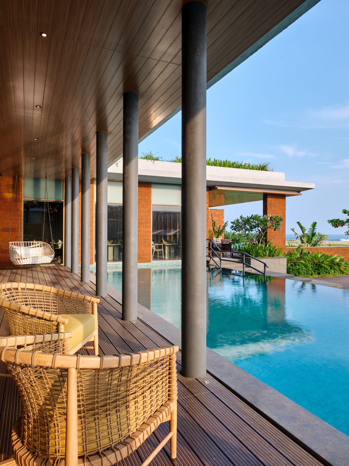 Covered patio with wicker chairs overlooks a serene infinity pool and lush garden. The setting is peaceful, with clear skies and modern architecture.