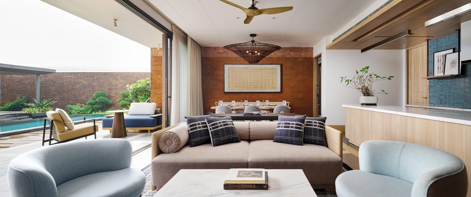 Modern presidential suite living room with a beige sofa and blue armchairs, opening to a patio with a pool. The space is airy and bright, with a peaceful ambiance.