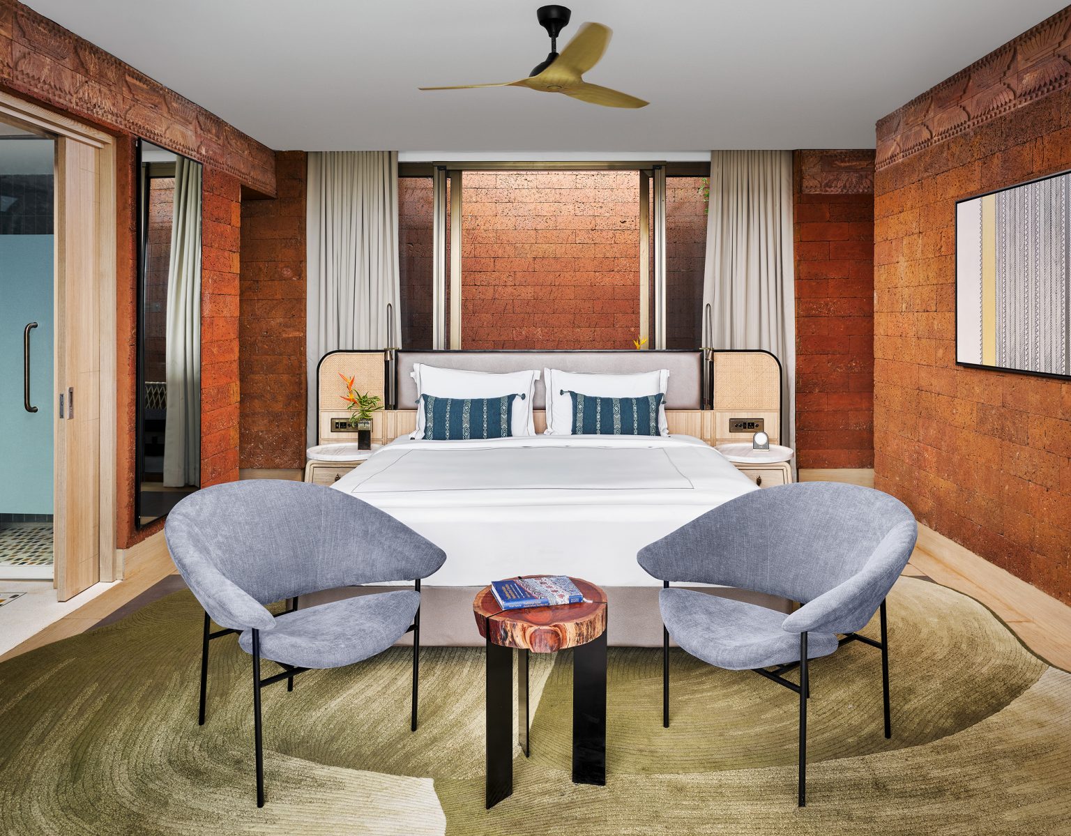Modern presidential suite bedroom with earth-toned walls, a white bed with blue cushions, and two gray chairs. A small wooden table and ceiling fan add warmth. Cozy and elegant.