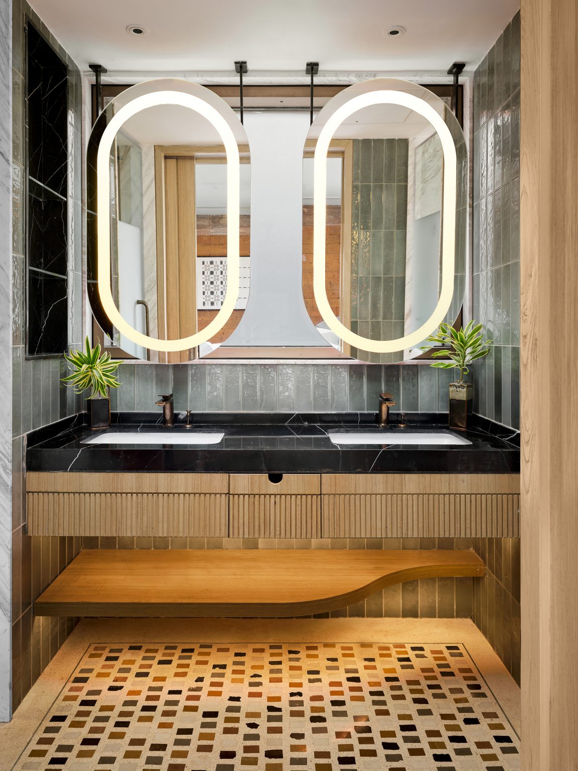 Modern bathroom with two oval backlit mirrors above a black marble double sink. Flanked by plants, a wooden shelf, and mosaic tile flooring.