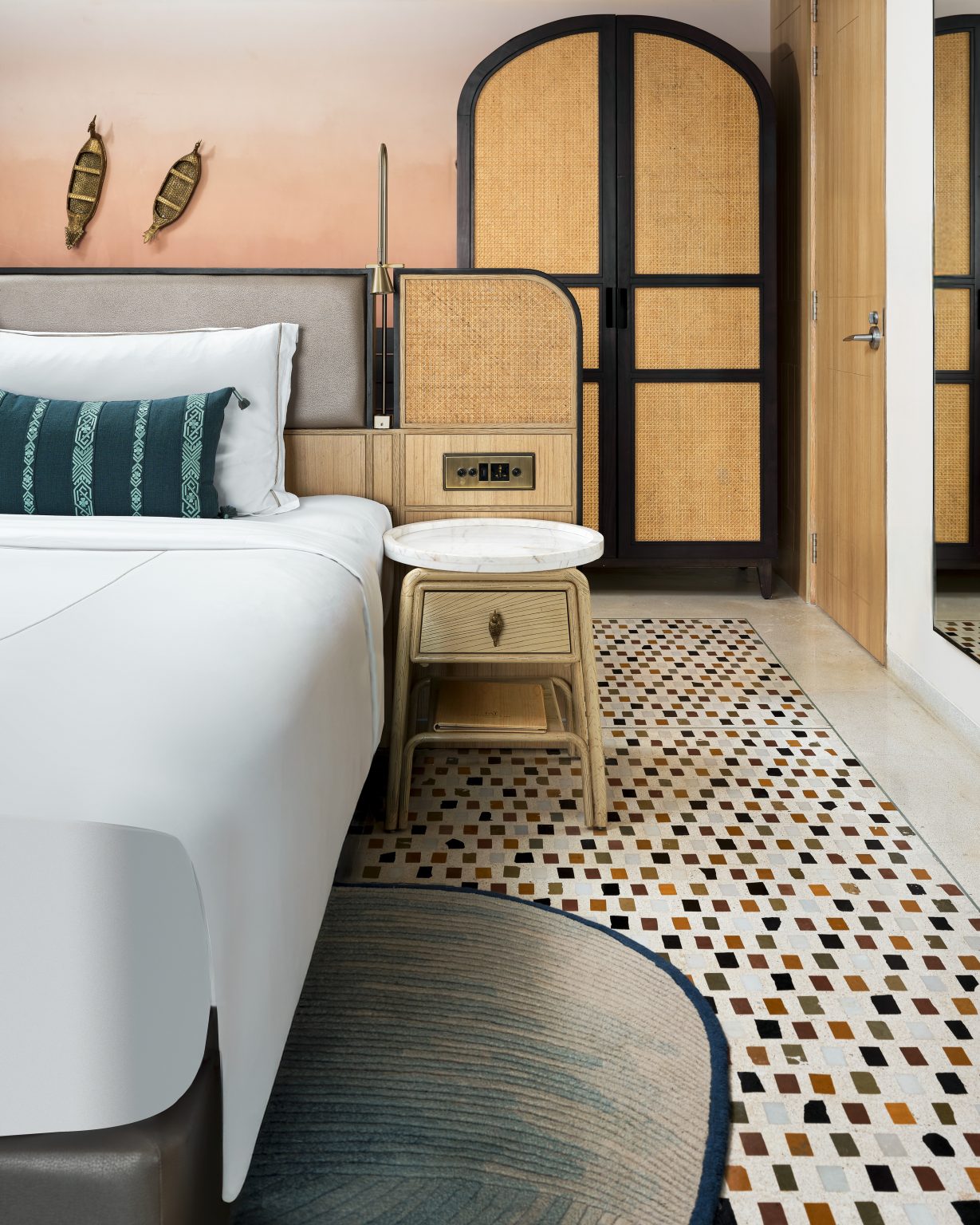 Stylish hotel room with a modern design. Features a white bed, patterned cushions, wicker wardrobe, round side table, and mosaic tiled floor. Cozy ambiance.