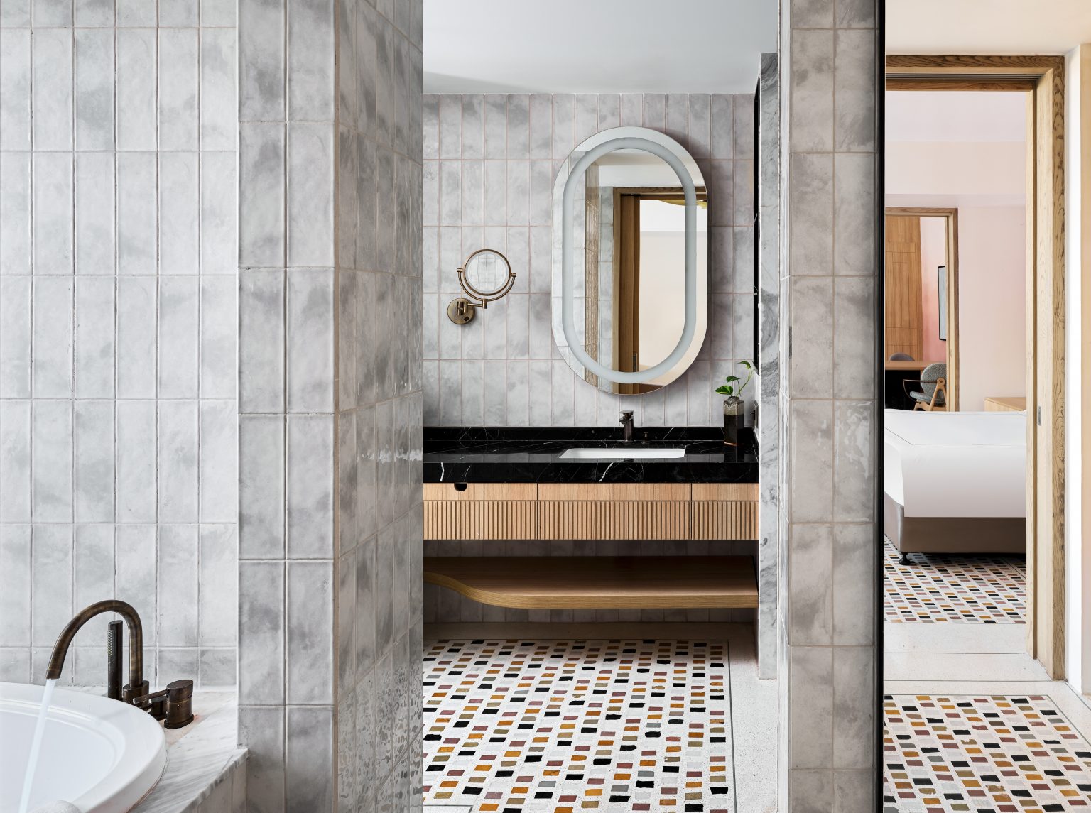 Modern bathroom with gray tiled walls, a black countertop, and wood accents. An oval mirror hangs above the sink. Mosaic floor adds color.