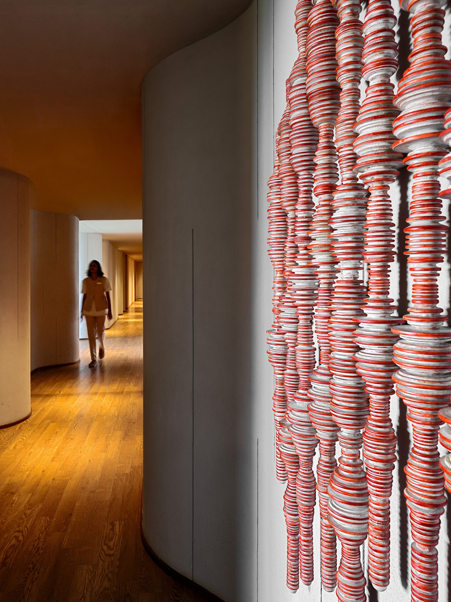Hallway with wooden floors and curved walls. A woman walks toward a vibrant, textured wall art made of red and white stacked discs, creating a dynamic pattern.