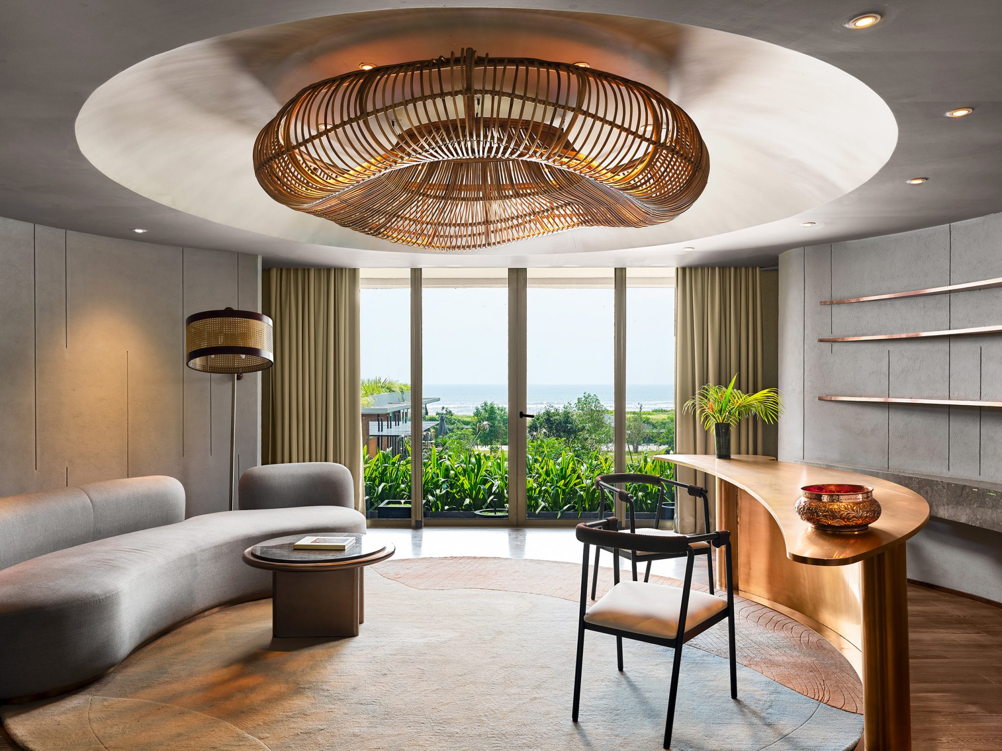 Modern living room with large windows showing a garden and sea view. Curved sofa, wooden coffee table, and bamboo ceiling light create a serene ambiance.