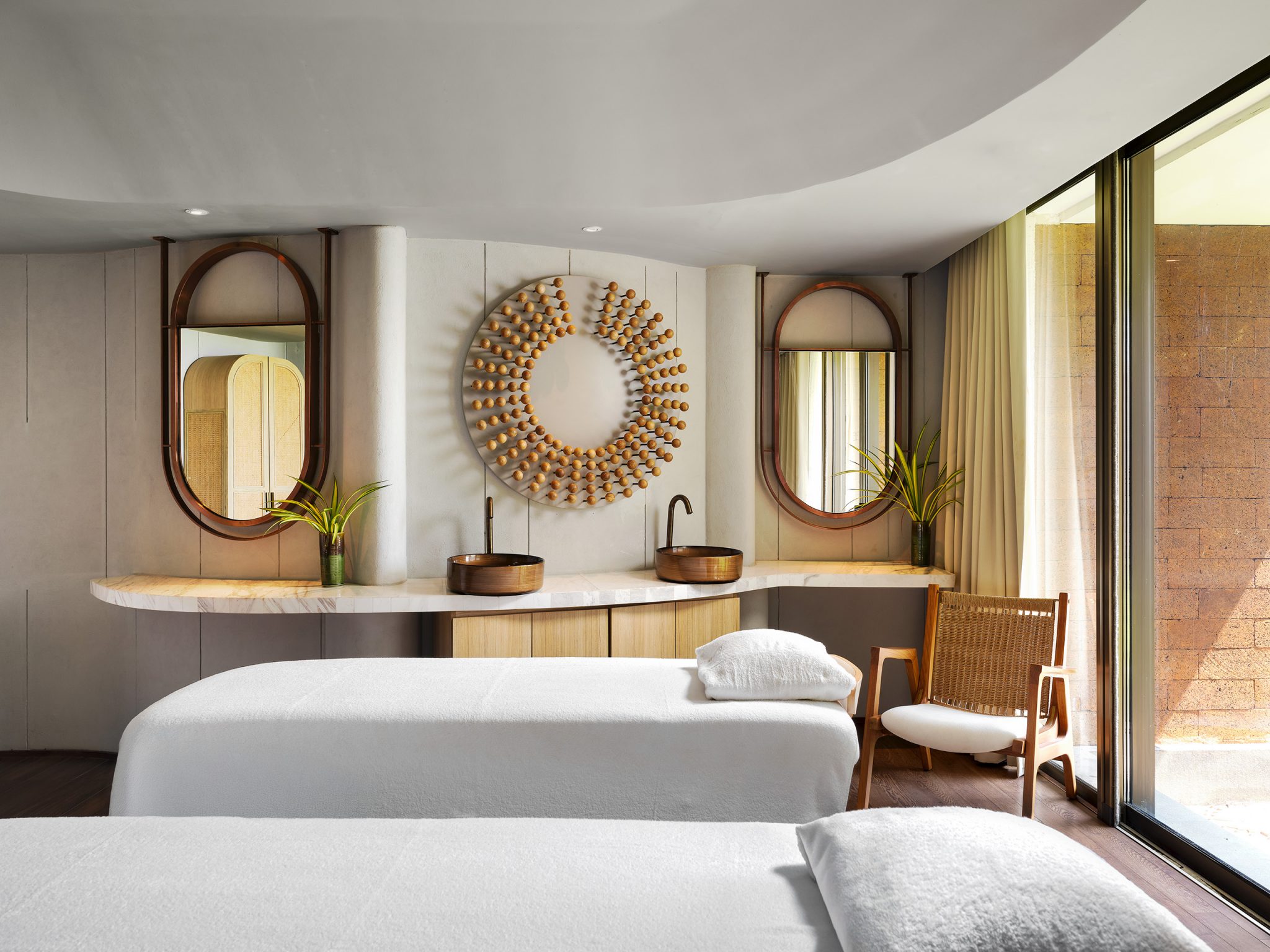 Modern spa room with two massage tables, a wicker chair, large circular mirrors, and a decorative sunburst centerpiece. Soft lighting and serene ambiance.