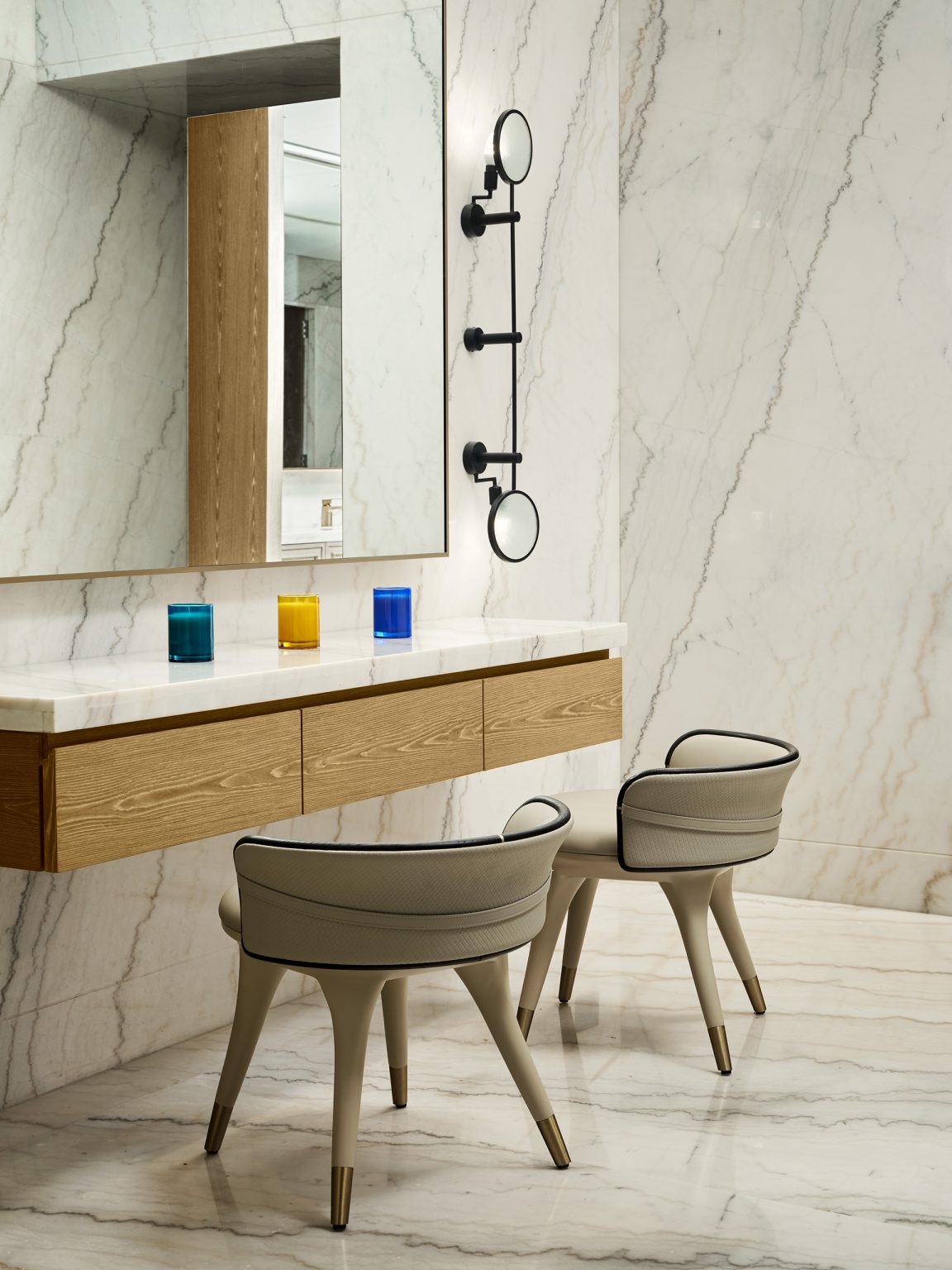 Elegant bathroom with marble walls and floor, wood vanity with white countertop, colorful cups, two modern beige chairs, and vertical mirrors.