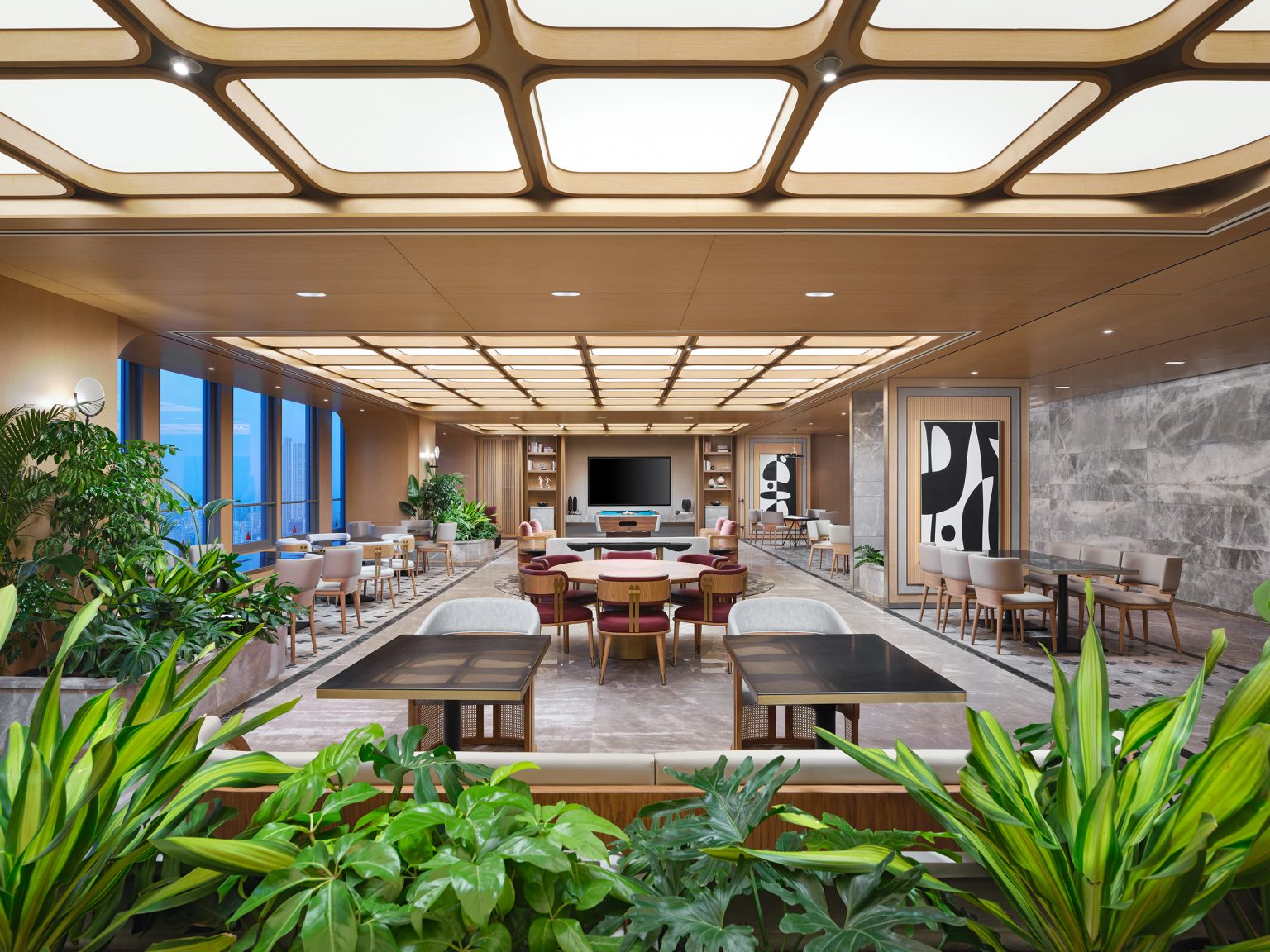 A modern, spacious lounge with a warm wood ceiling, geometric lighting, vibrant plants, and abstract art. It features cozy seating and large windows, creating an inviting ambiance.
