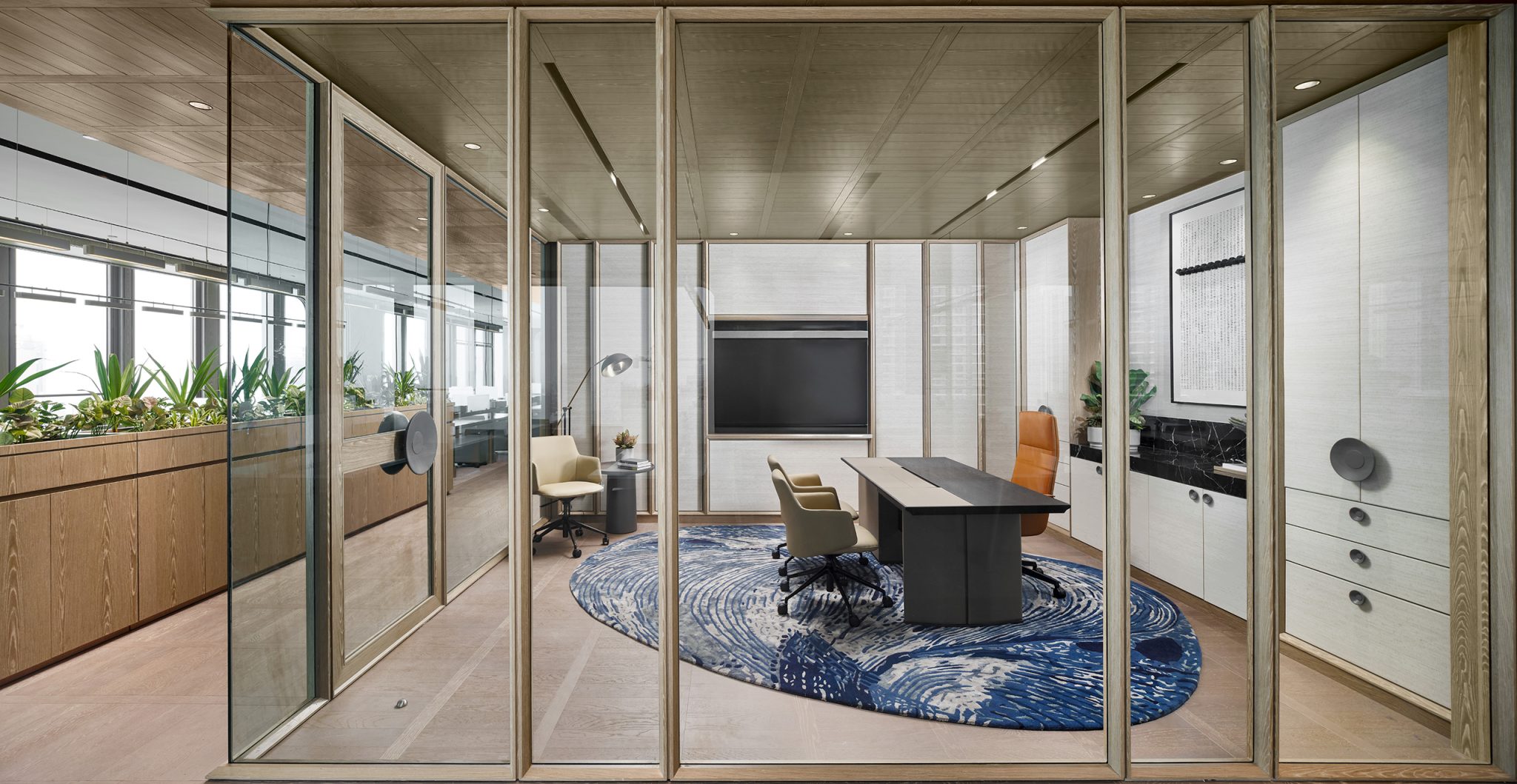 Modern office executive cabin with glass walls, wooden accents, and a large desk on a blue wave-patterned rug. Neutral tones and plants create a calm, professional atmosphere.