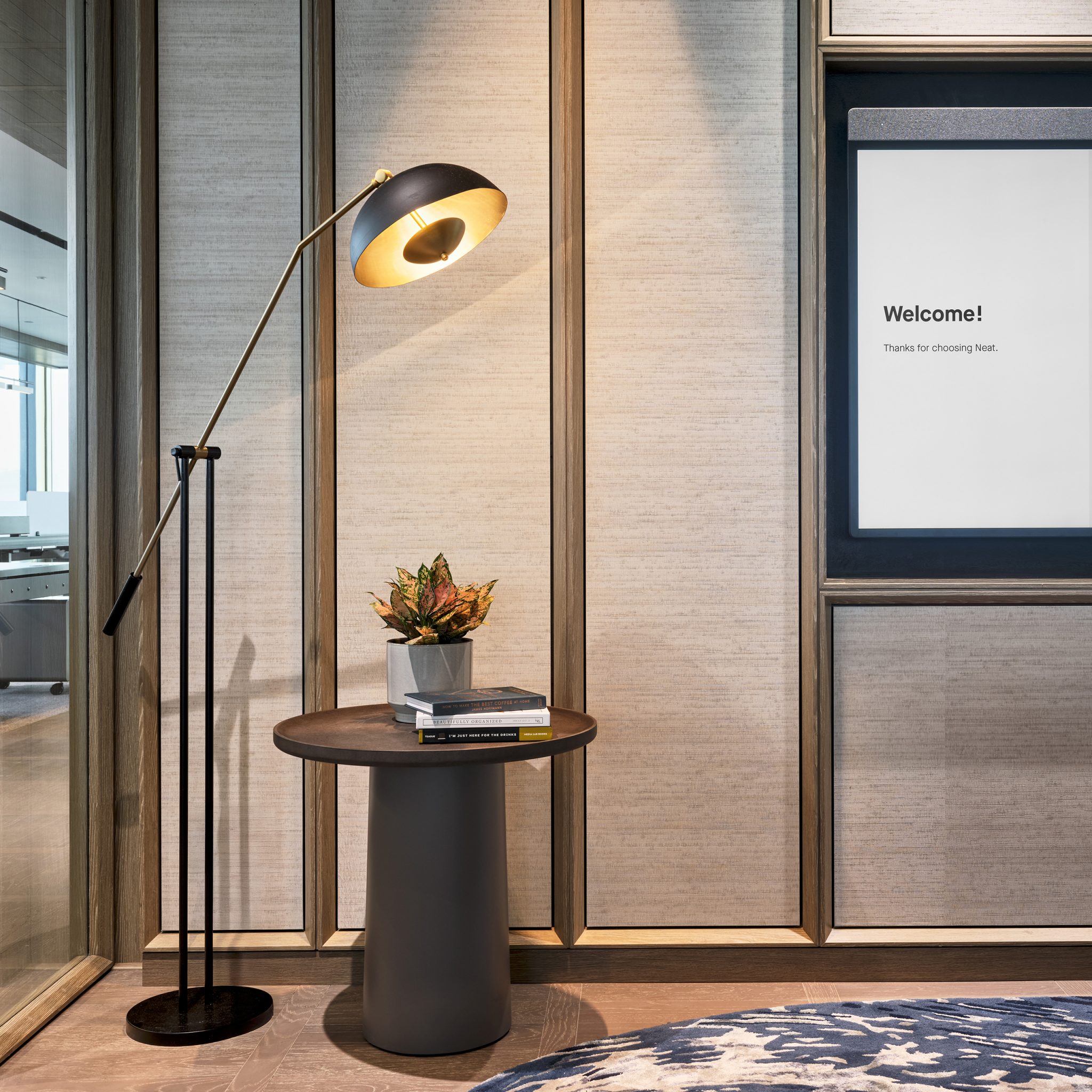 A modern interior corner features a sleek floor lamp beside a round table with a small plant and books. A screen on the wall displays 