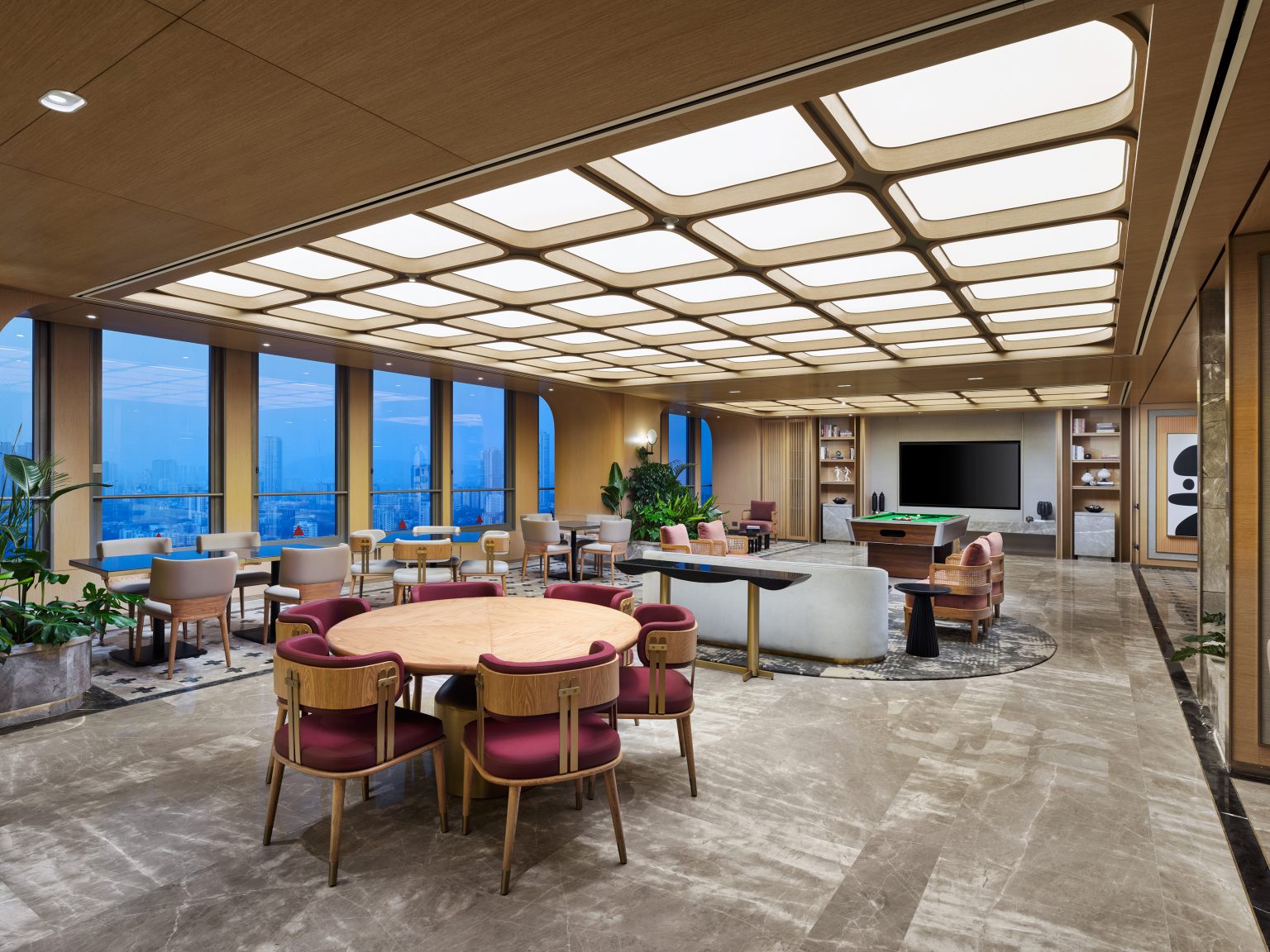 Spacious lounge with modern decor featuring large windows with city view, round tables with red chairs, a white sofa, and a wall-mounted TV.