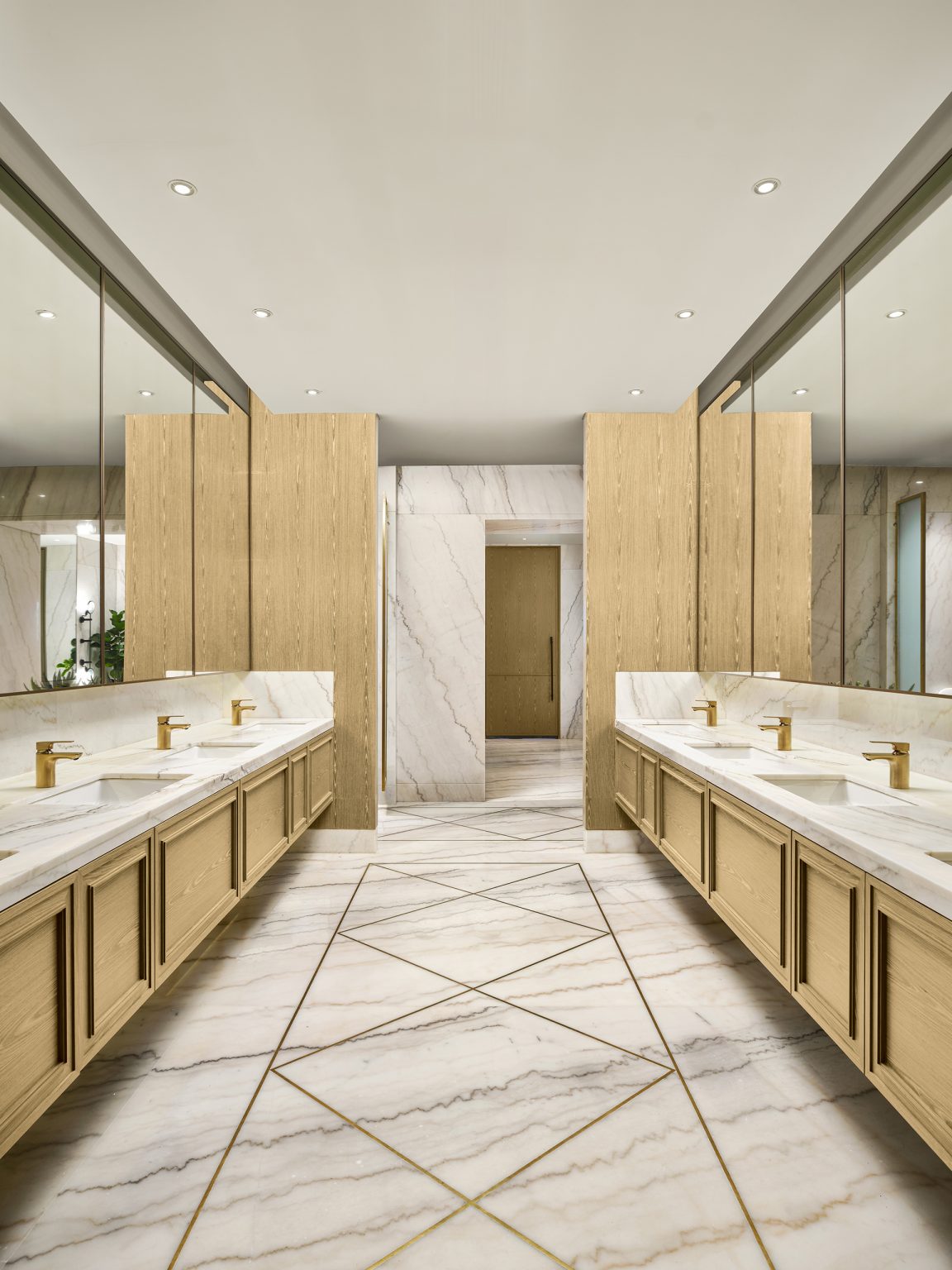 Luxurious restroom with marble floors and counters, two long vanities, gold fixtures, and wooden accents. Mirrors and soft lighting create an elegant ambiance.