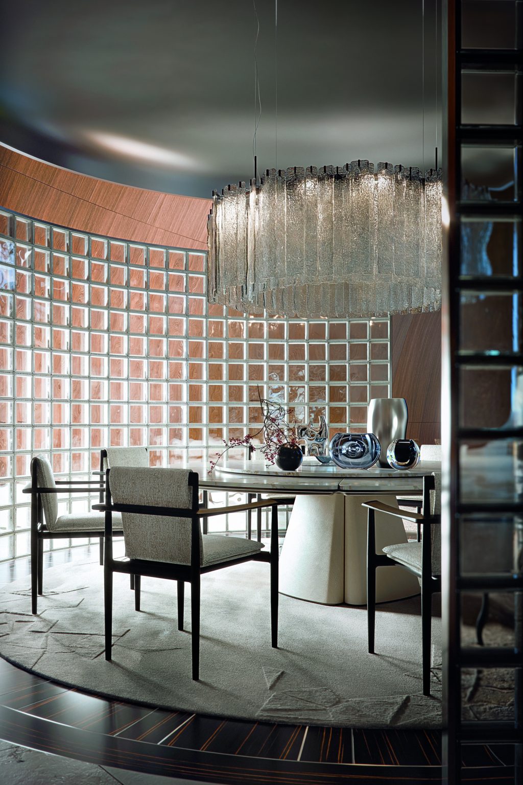 Elegant dining room featuring a round table with Giorgetti Kumiki chairs, a decorative centerpiece, and a large, textured chandelier. A wall of glass blocks creates a soft, ambient light, enhancing the room's sophisticated atmosphere.
