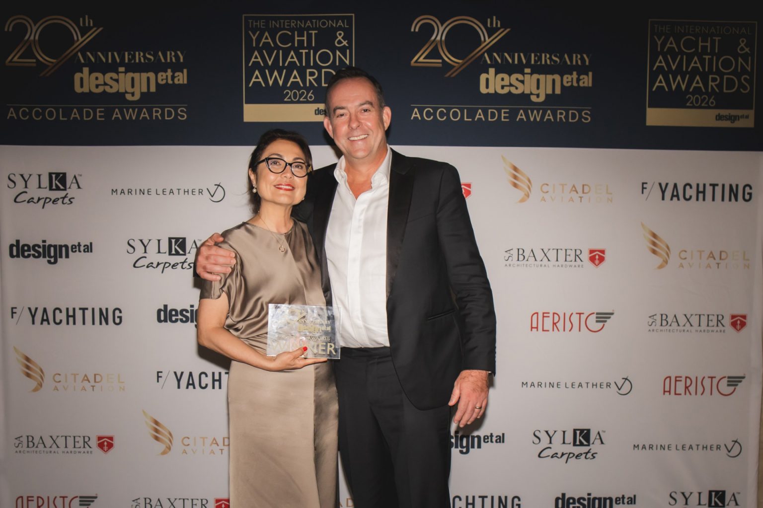 A smiling man and woman pose together at the Yacht & Aviation Awards. The woman holds an award. The backdrop features event logos and sponsor names.