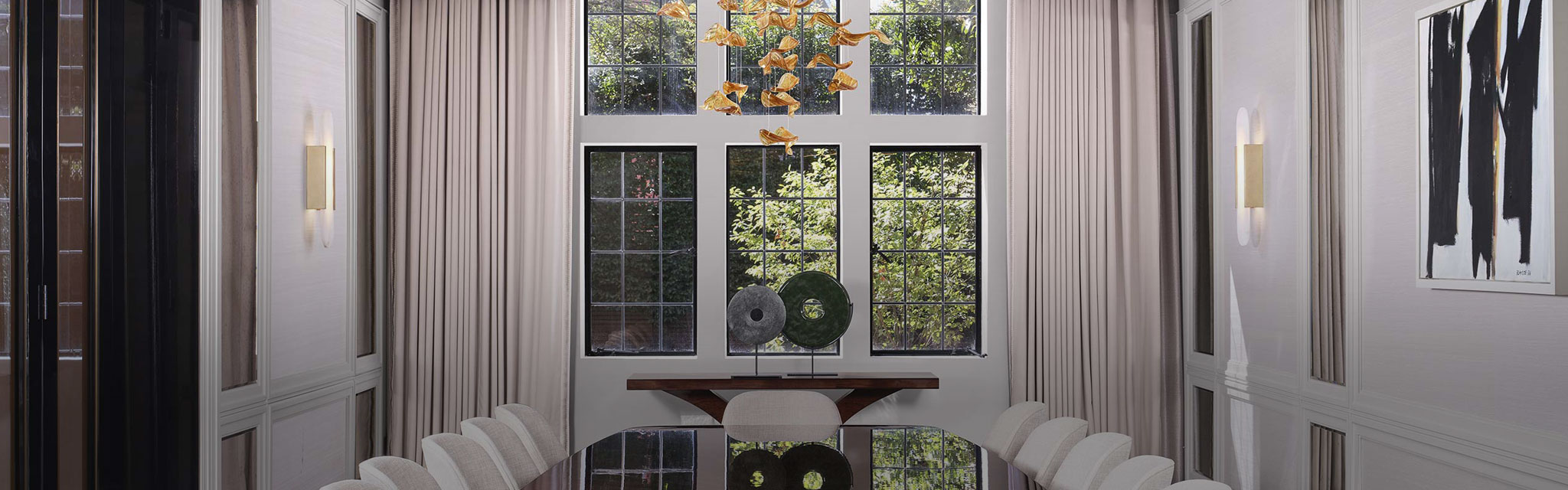 Elegant dining room with a glossy black table, white chairs, and a striking gold chandelier. Large windows reveal lush greenery outside. Sophisticated ambiance.