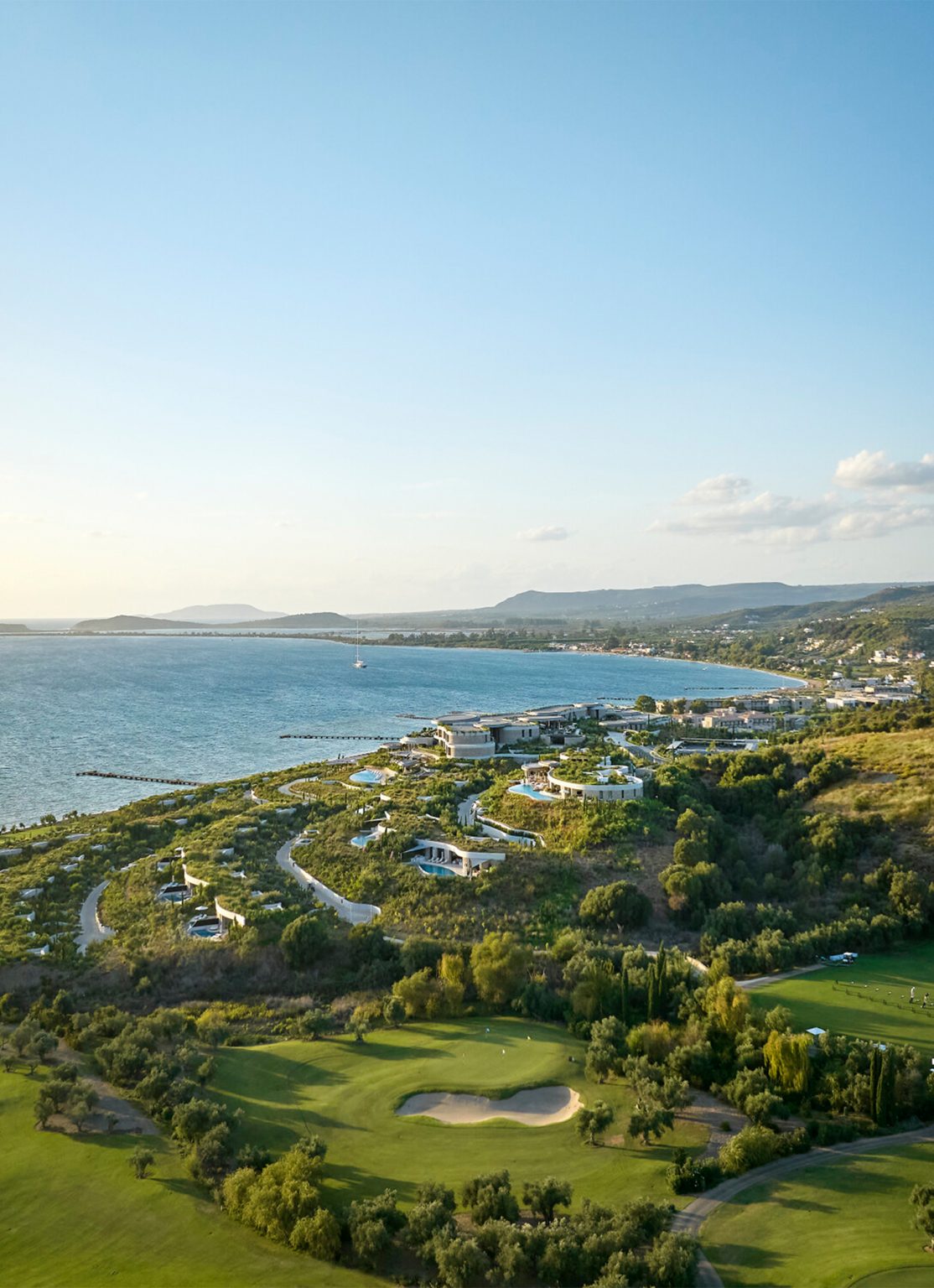 Aerial view of a luxury coastal resort in Greece with grass-roofed buildings, a golf course, and an ocean bay view.