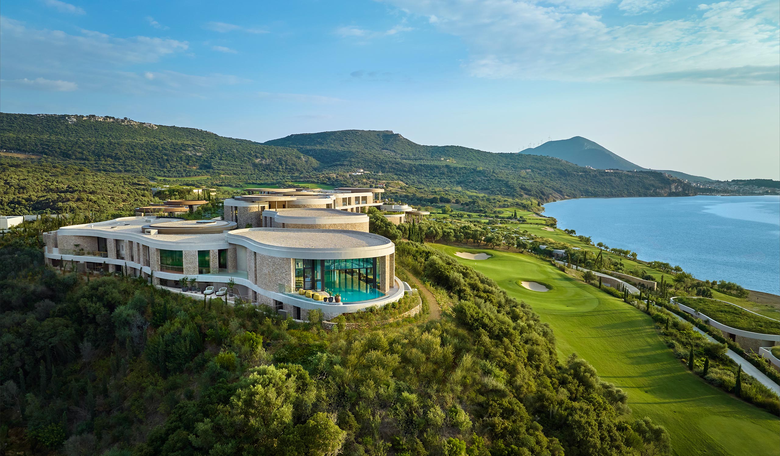 Modern, luxurious hilltop mansion with floor-to-ceiling windows overlooking lush greens and a serene coastline. The atmosphere is tranquil and scenic.