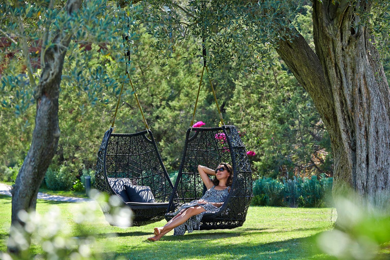 A woman in a sundress and sunglasses relaxes in a hanging chair under a large tree on a sunny day. Lush greenery surrounds the peaceful scene.