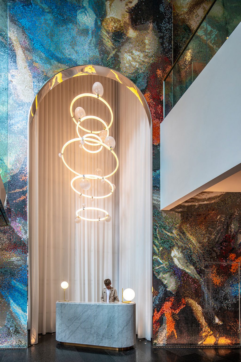 Grand hotel lobby featuring a multicolored mosaic wall, elegant spiral chandelier, and marble reception desk. A person is standing behind the desk.