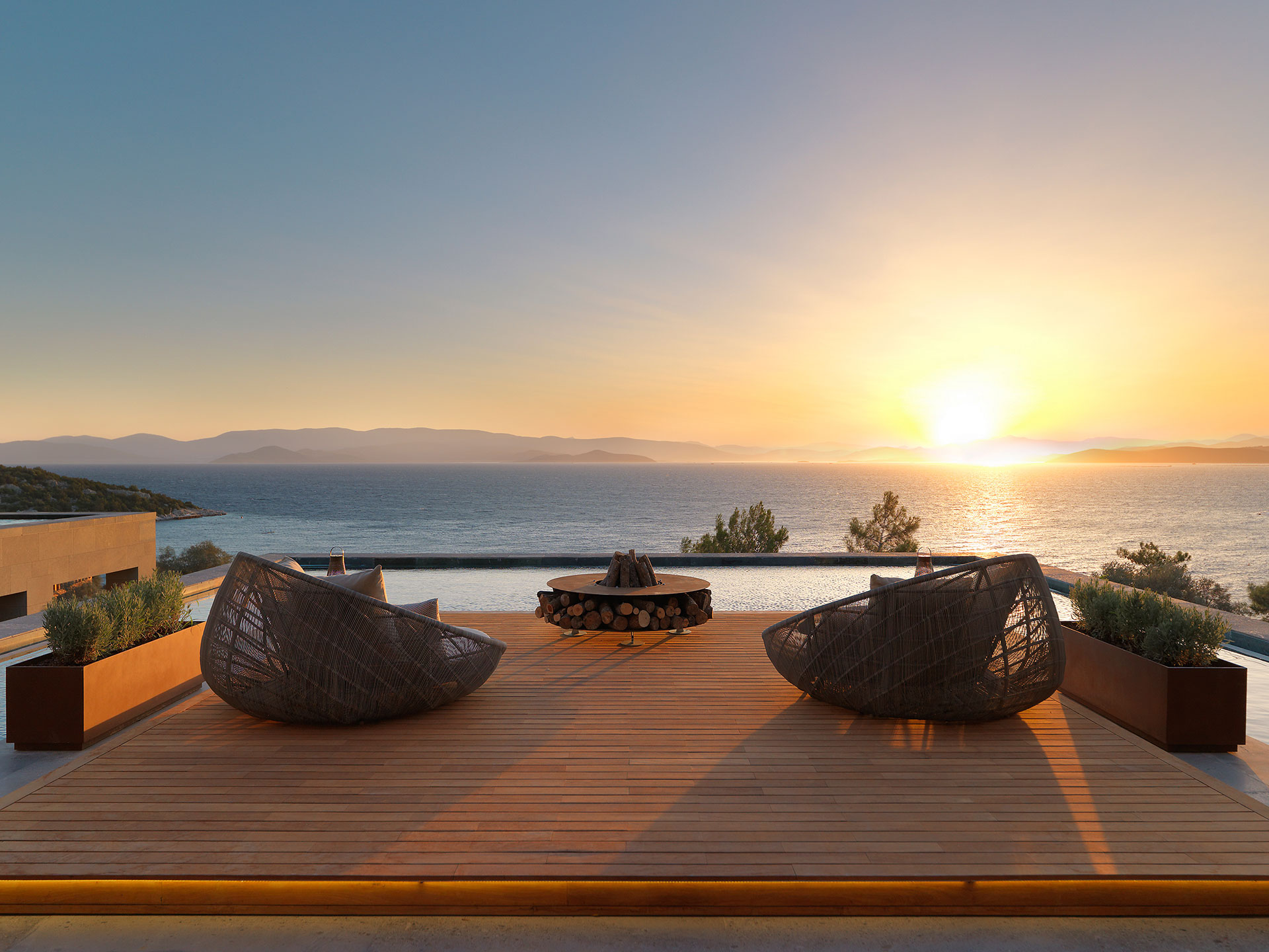 Modern patio with wicker chairs and a fire pit, overlooking a serene sea at sunset. Warm tones create a peaceful, relaxing ambiance.