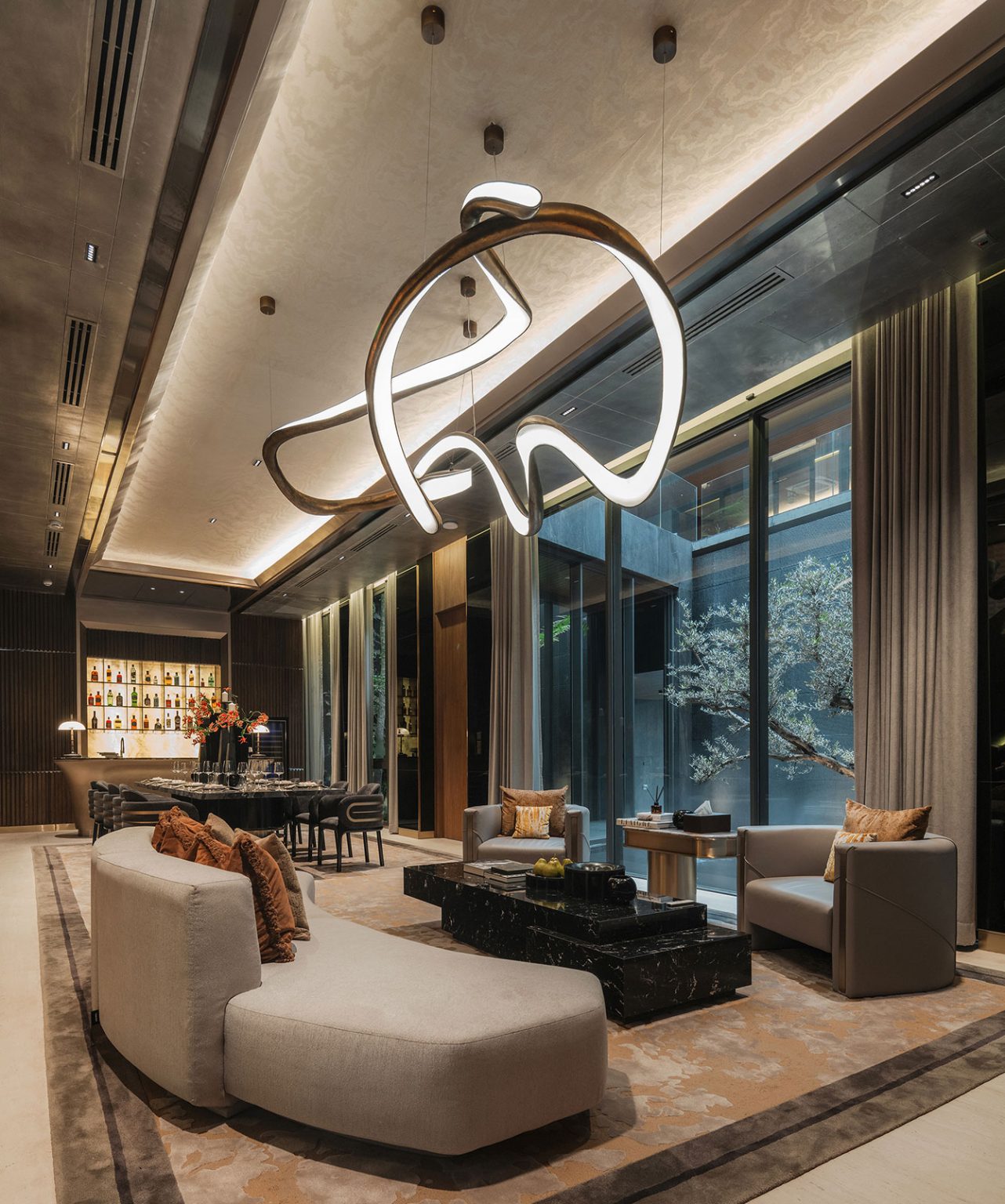 Luxurious living room with modern, abstract chandelier, beige curved sofa, and elegant armchairs. Large windows reveal serene outdoor view. Open-plan living room and dining area.