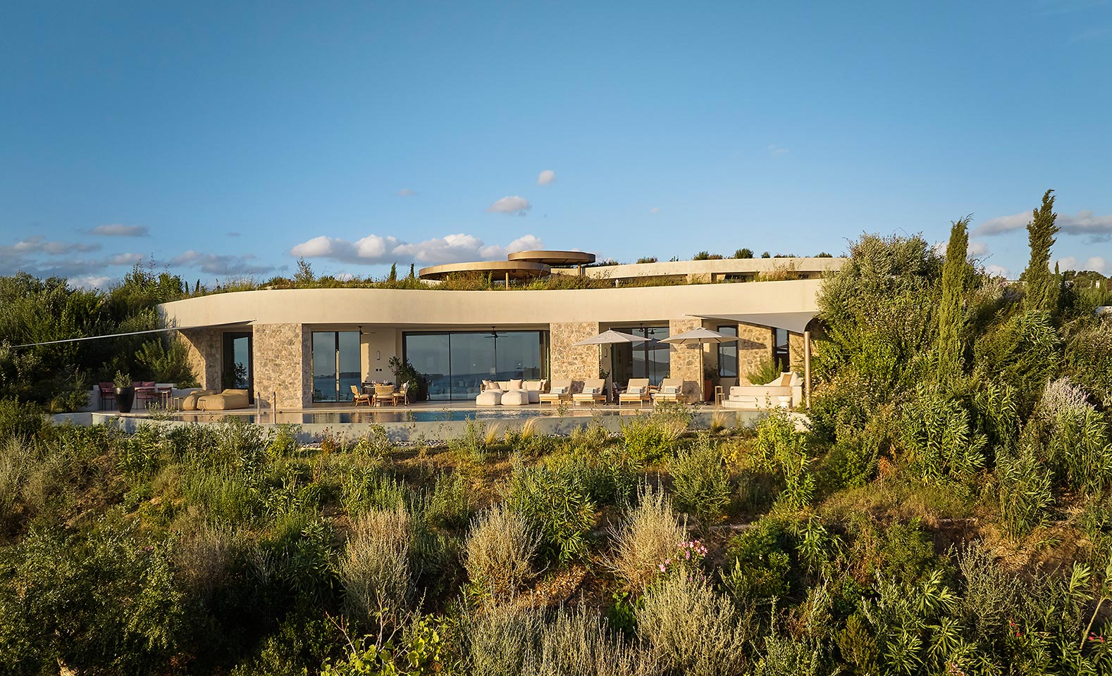 Modern villa with large glass windows, surrounded by lush greenery and shrubs. The sky is clear and blue, creating a serene, luxurious atmosphere.