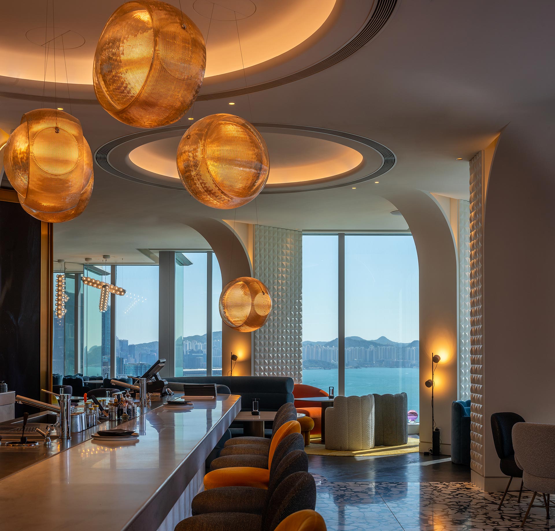 Modern bar interior with warm, glowing pendant lights, patterned decor, and sleek furniture. Large windows reveal a scenic city and lake view.