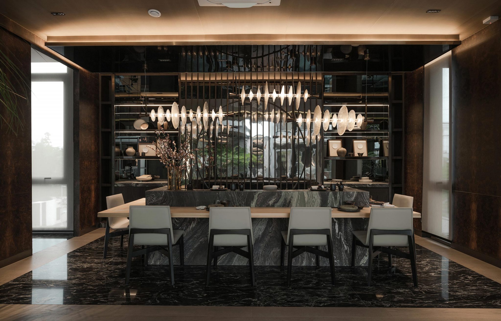 Luxurious dining room features a long marble table with six chairs, modern chandelier, shelving with decor, warm ambient lighting, and dark tones.