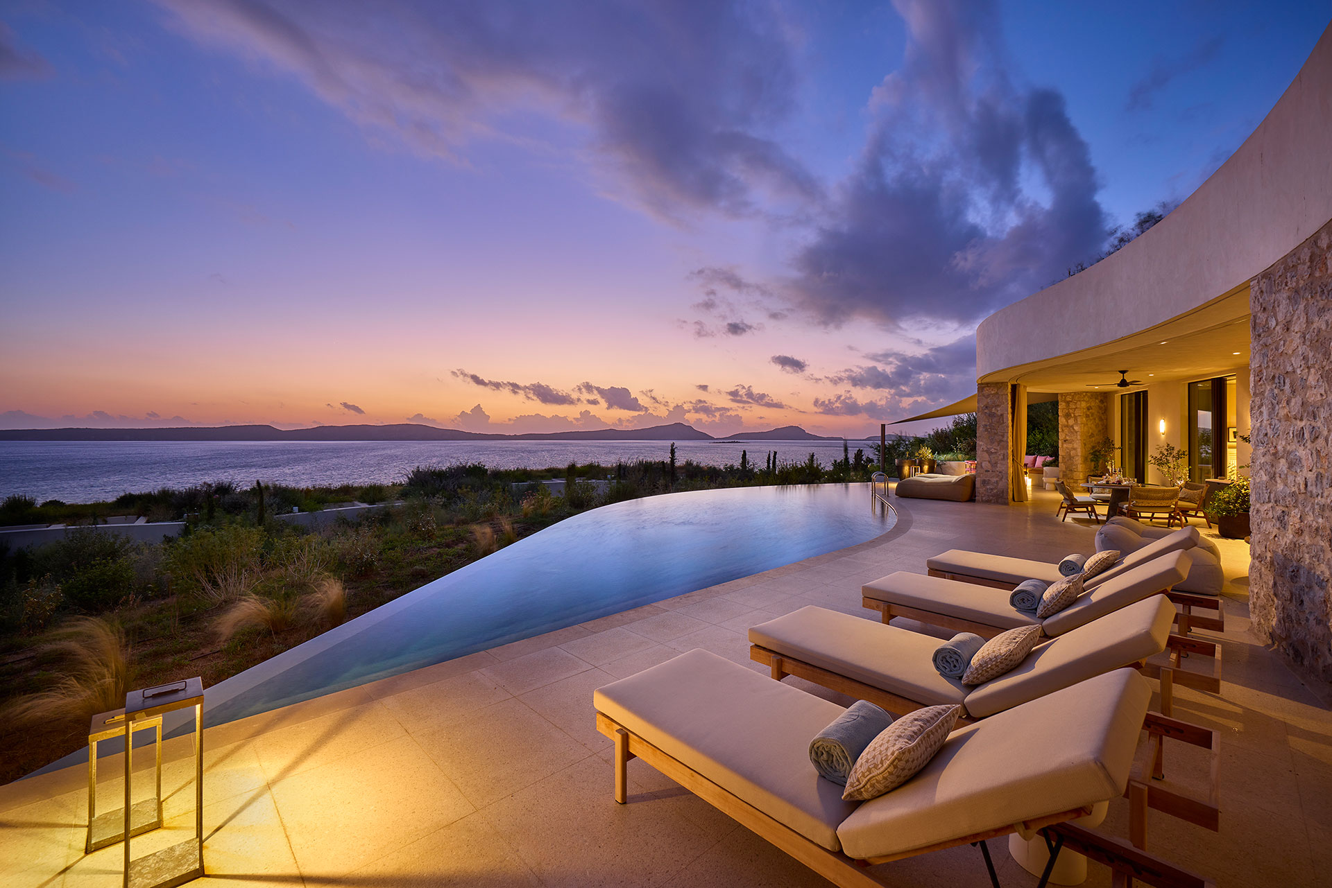 Luxurious poolside sea view at sunset featuring plush loungers, an infinity pool, and open seating area. Warm lighting enhances the serene ocean backdrop.