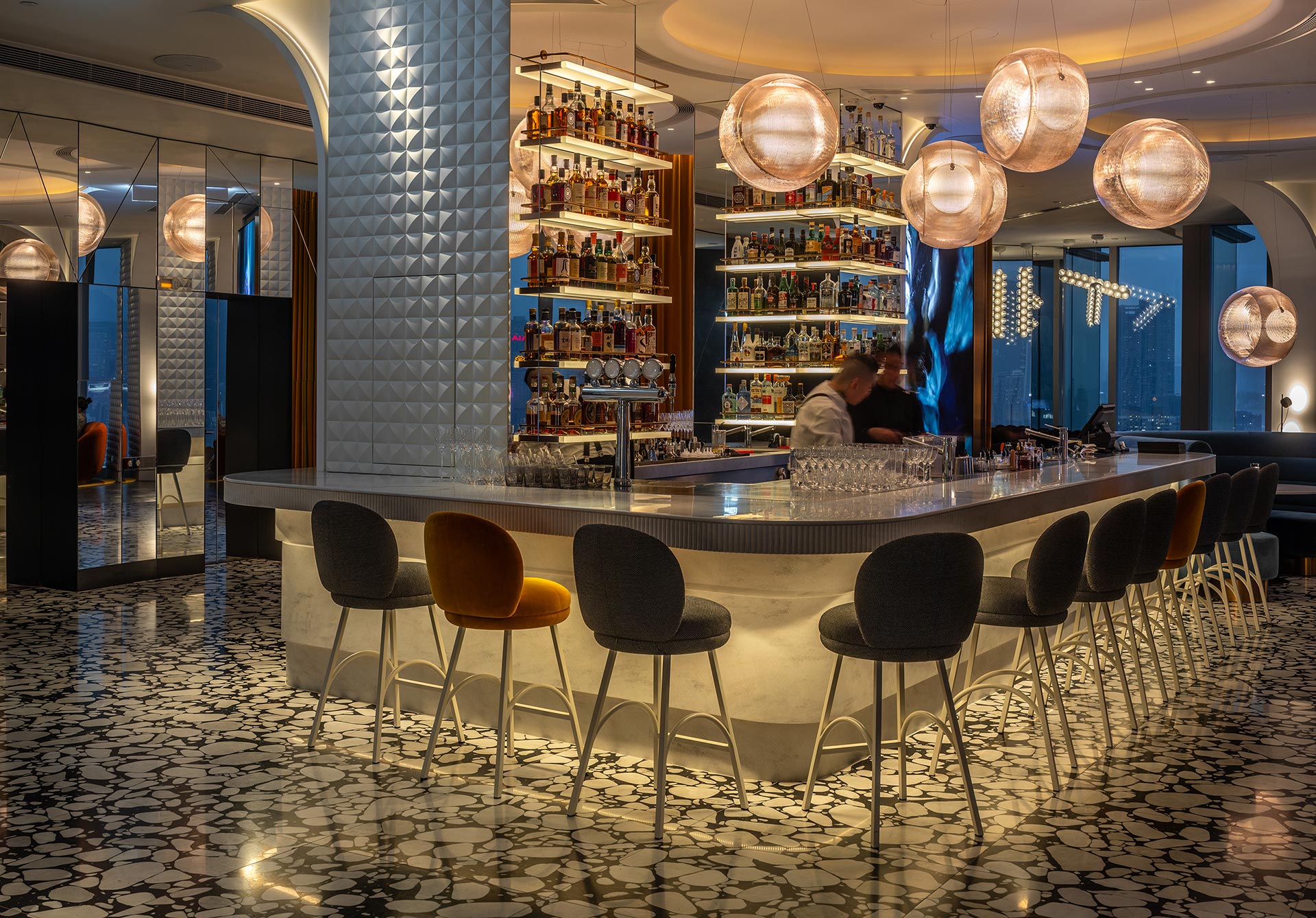 Sleek, modern bar interior with a long, curved marble counter surrounded by stylish chairs. Warm lighting from spherical lamps creates an inviting ambiance.