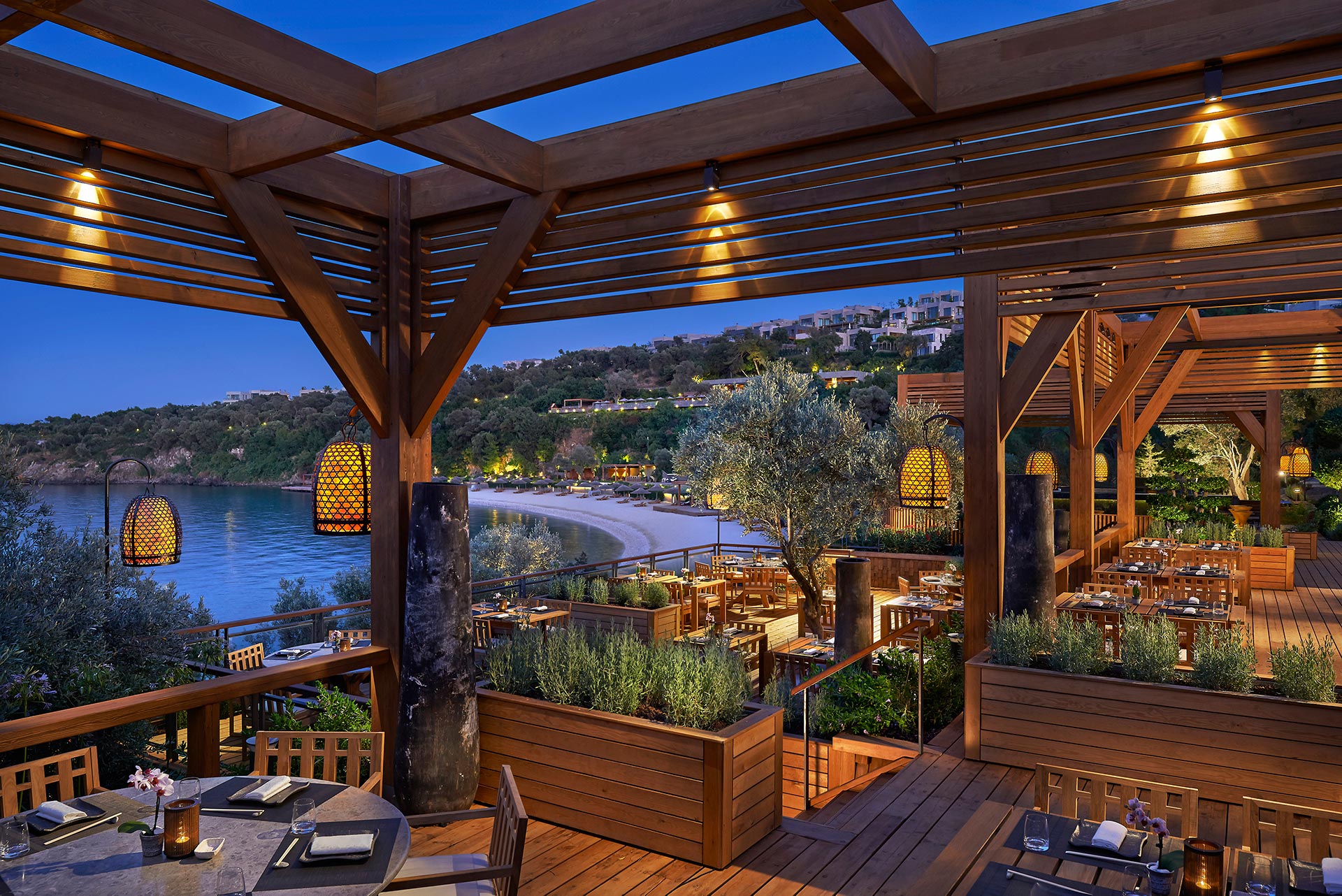 Scenic outdoor restaurant with wooden decor overlooking a serene bay. Warm lights and lush greenery create a cozy, inviting evening ambiance.